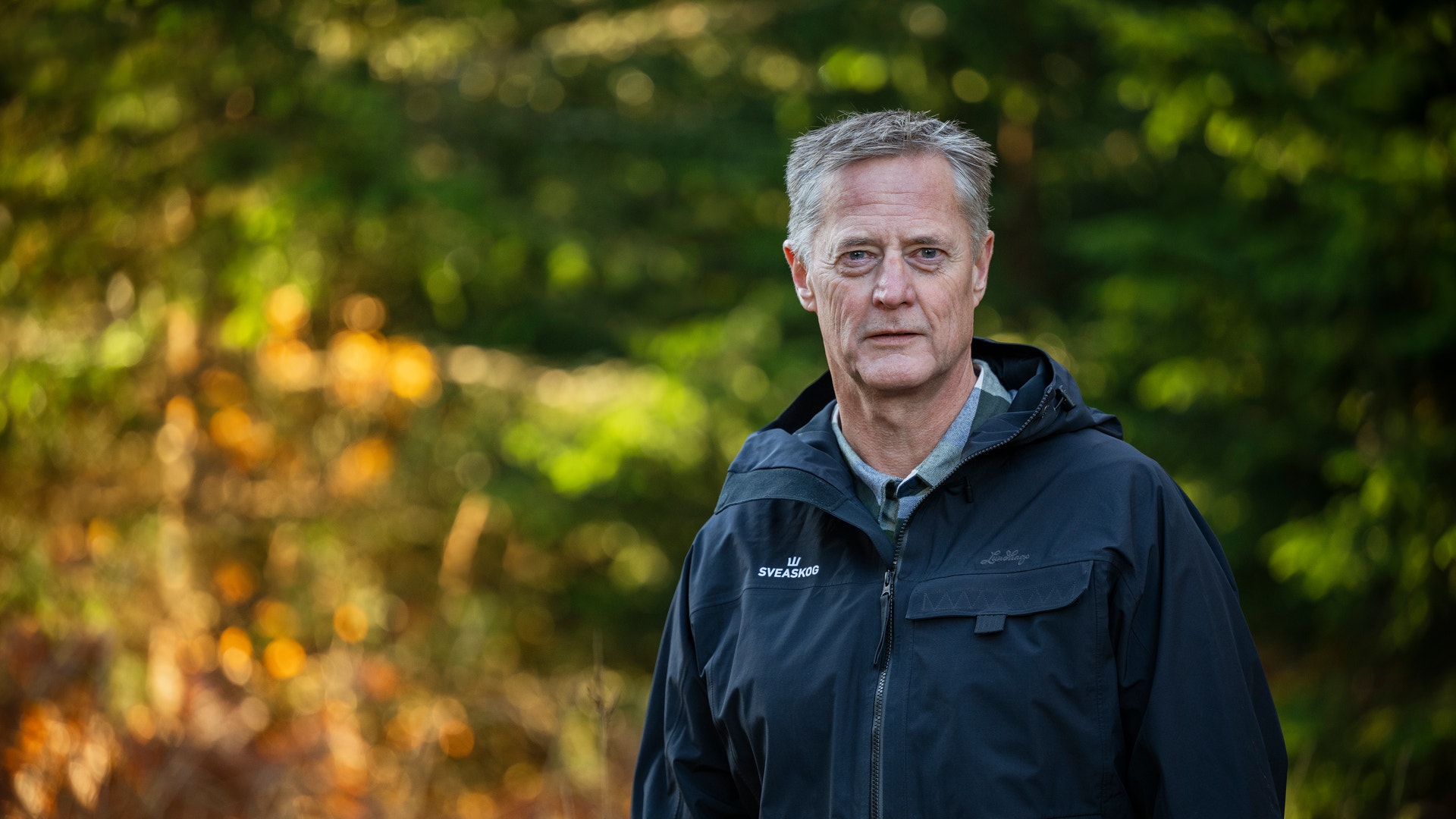 Porträtt av en äldre man med grått hår i mörk jacka som står utomhus framför skog i höstfärger.
