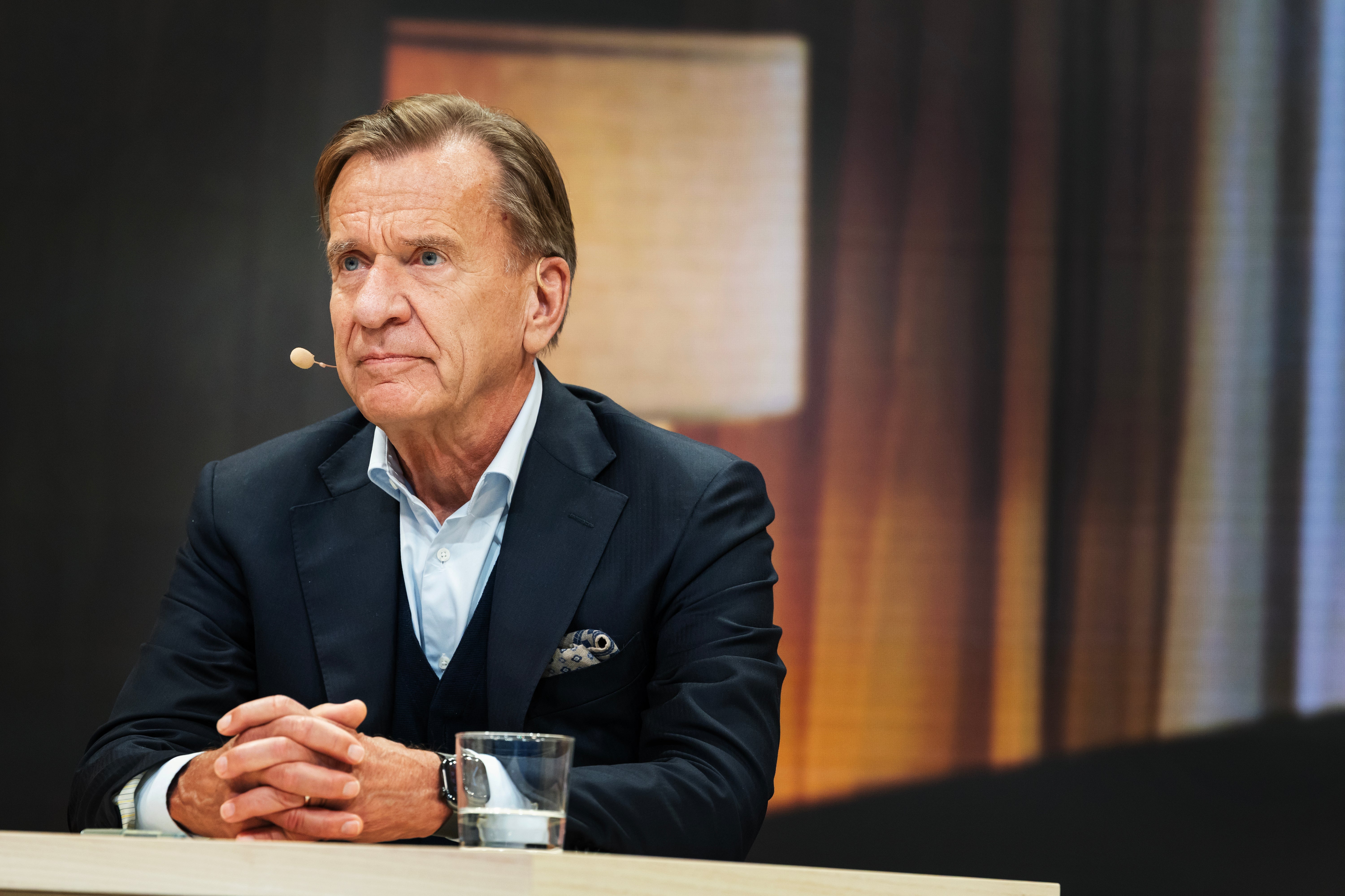 En man i kostym med mikrofon sitter vid ett bord under en intervju eller paneldiskussion.