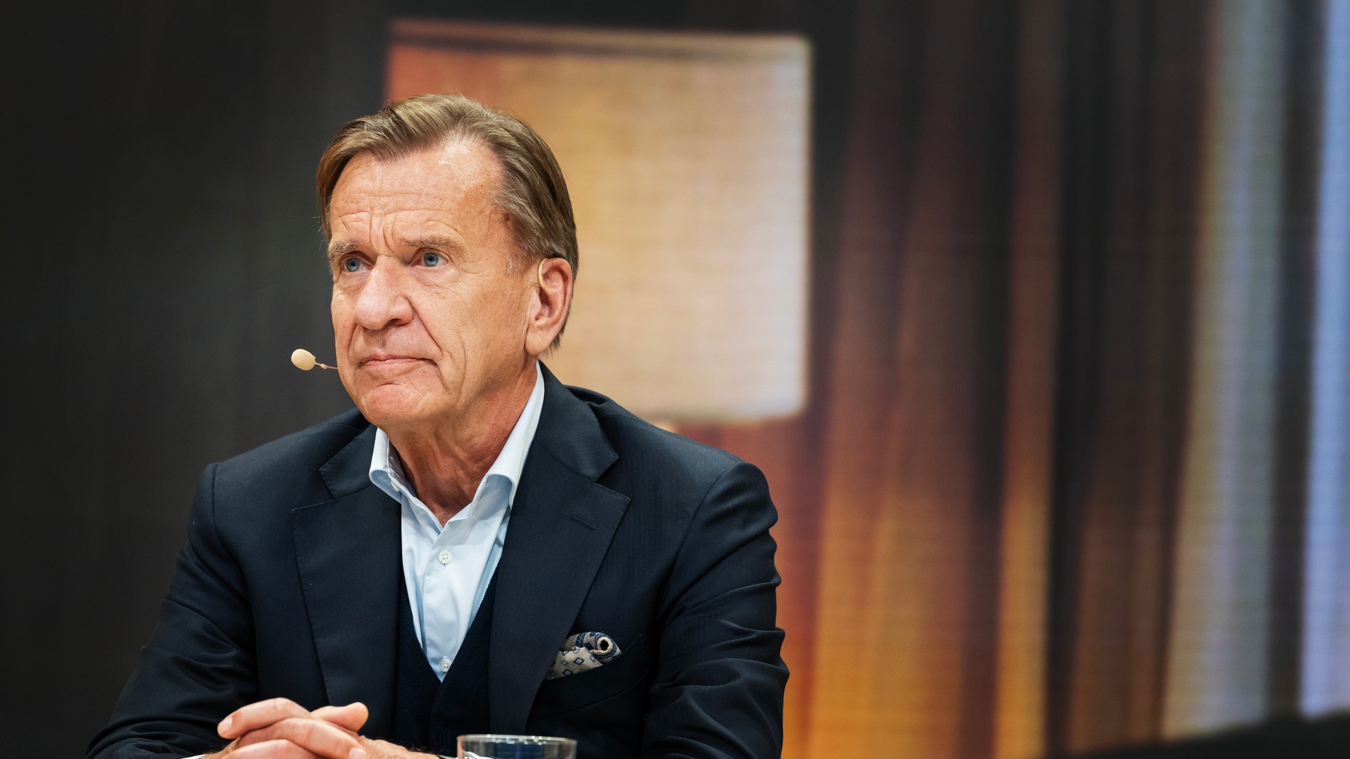 En man i kostym med mikrofon sitter vid ett bord under en intervju eller paneldiskussion.