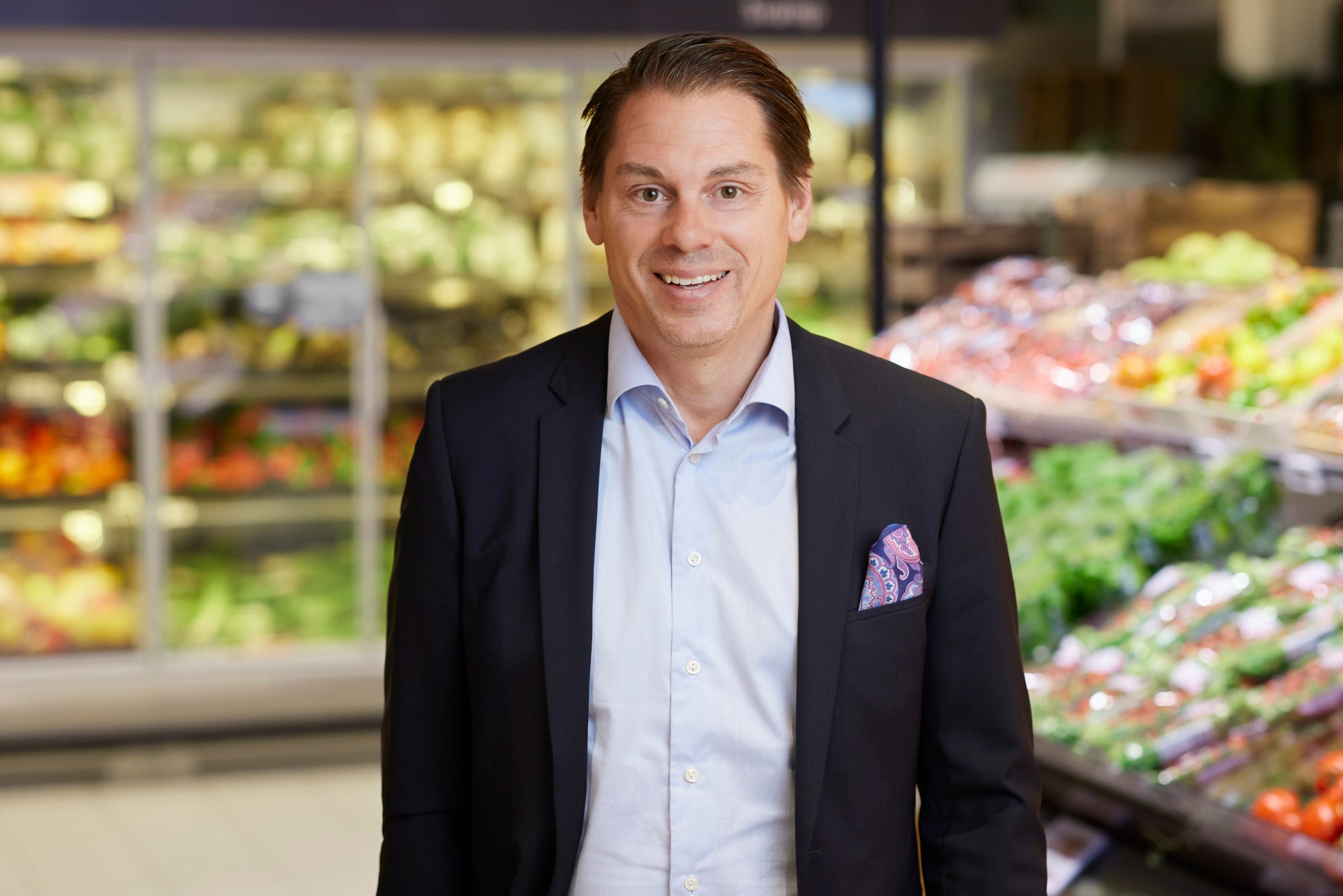 En man i kostym ler mot kameran, stående i en butik omgiven av frukt- och grönsakshyllor.