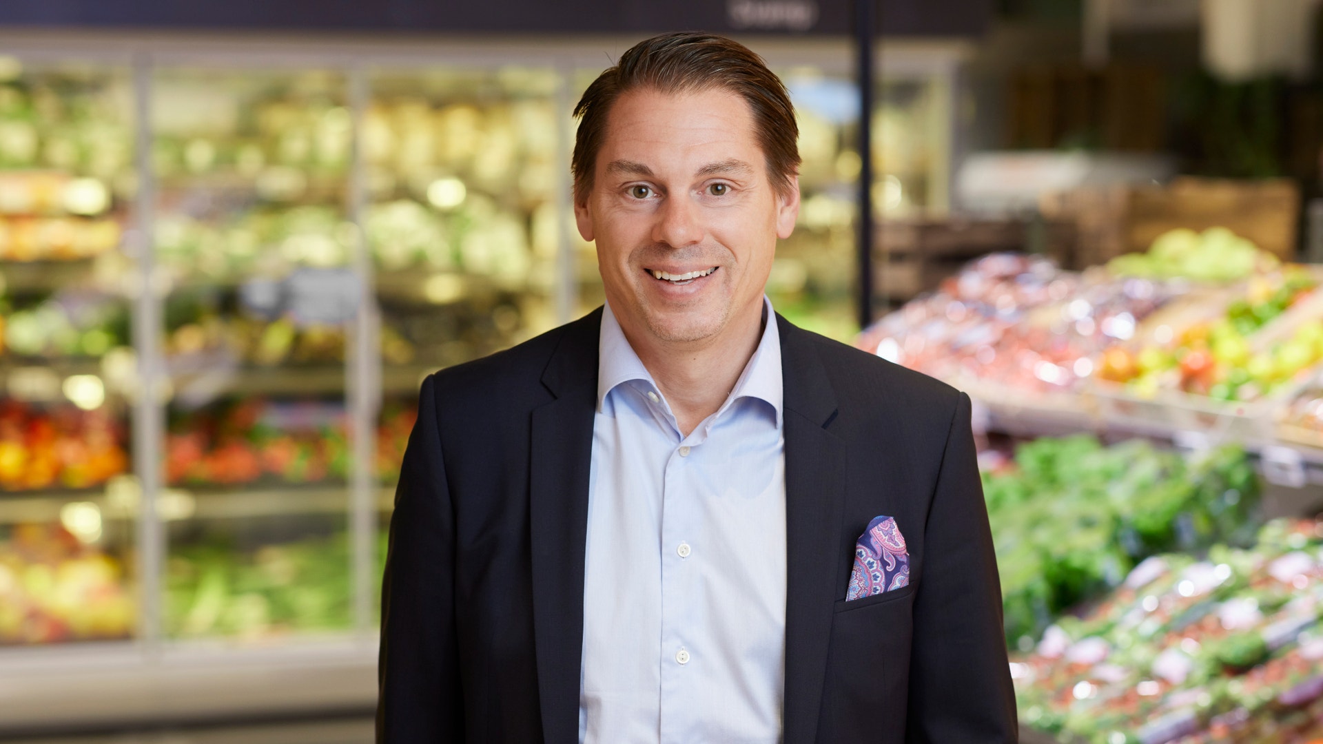 En man i kostym ler mot kameran, stående i en butik omgiven av frukt- och grönsakshyllor.