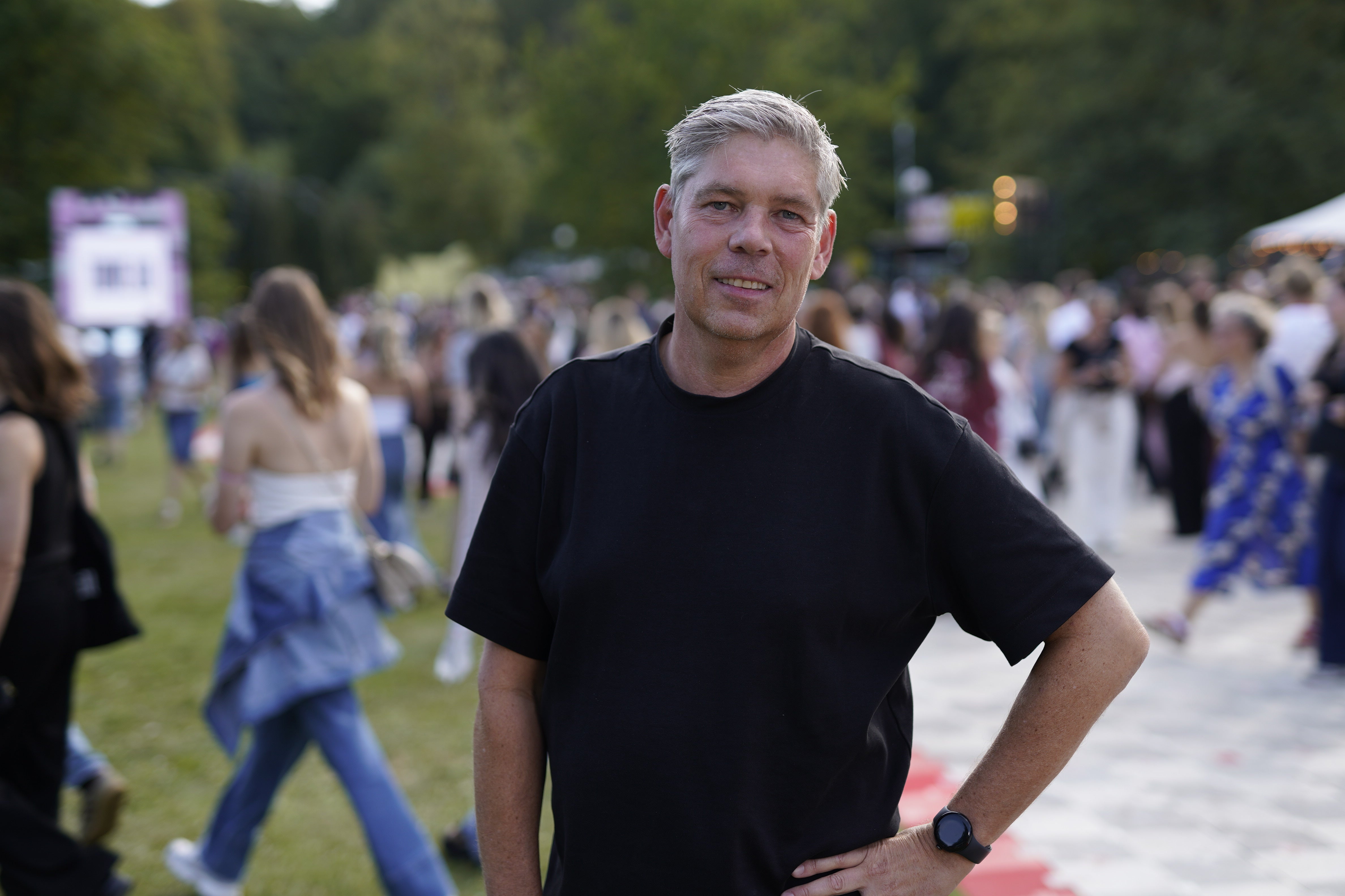En man i svart t-shirt står på en utomhusevent och ler mot kameran. I bakgrunden syns många människor på en gräsmatta.