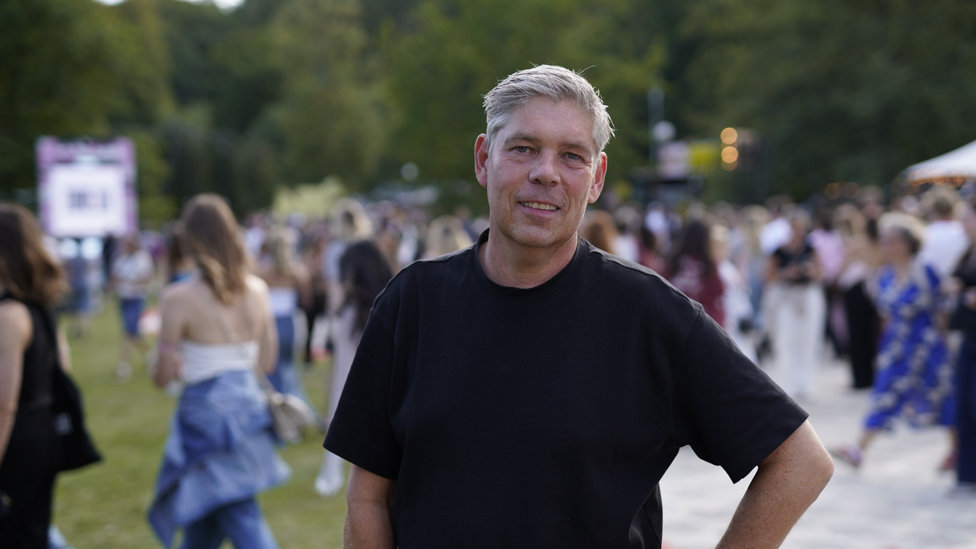 En man i svart t-shirt står på en utomhusevent och ler mot kameran. I bakgrunden syns många människor på en gräsmatta.