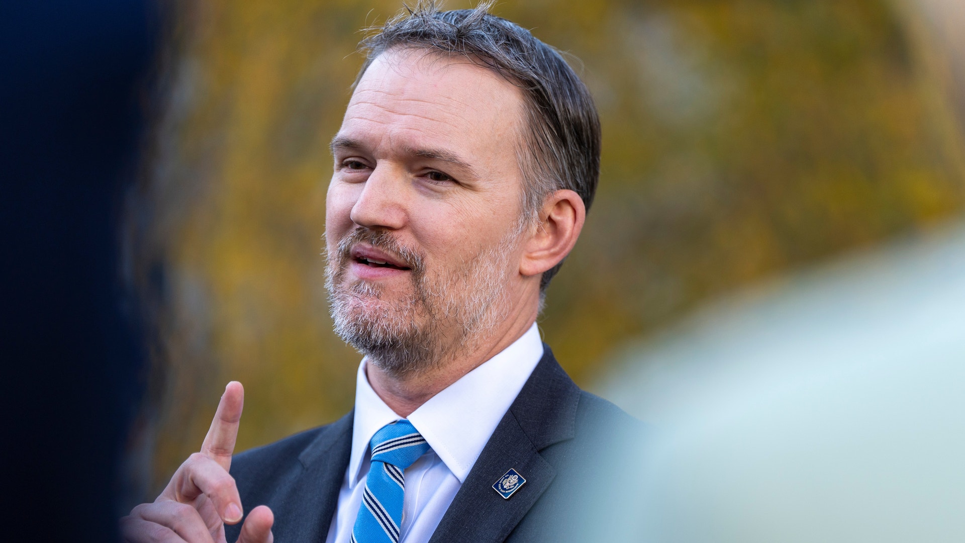 Man i mörkblå kostym och blå slips som gestikulerar under ett tal. Höstfärgad bakgrund.