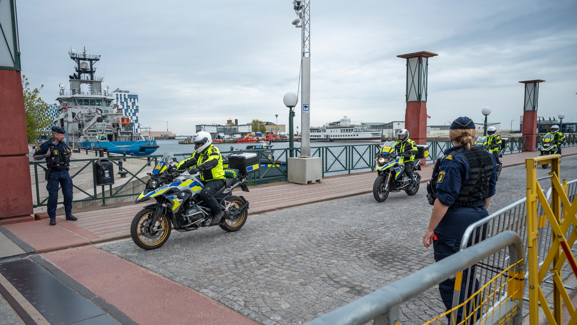 Polismotorcyklister i gul och blå utrustning utför en demonstration på en hamn. Båtar och hamnindustri syns i bakgrunden.