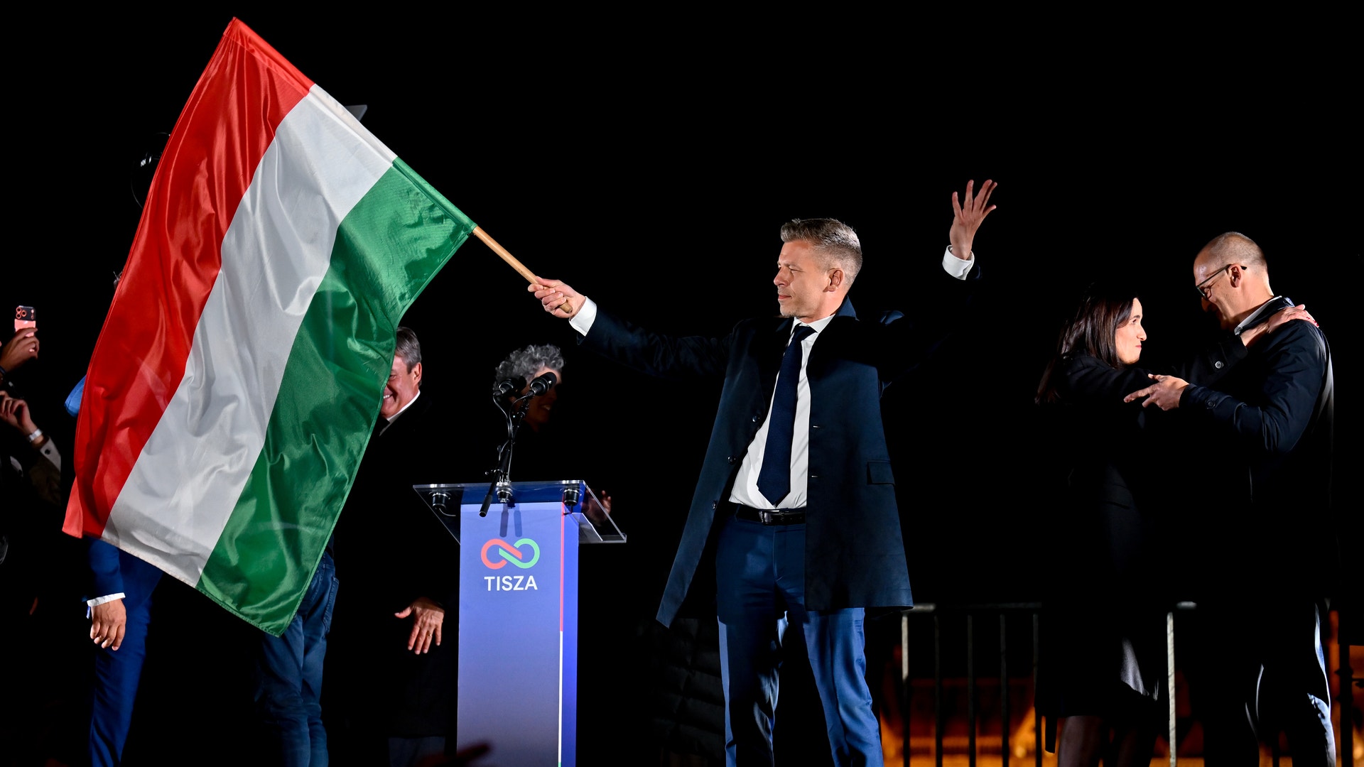 En man i kostym talar vid ett podium under en politisk manifestation. En ungersk flagga hålls upp i bakgrunden och flera personer står omkring på scenen.