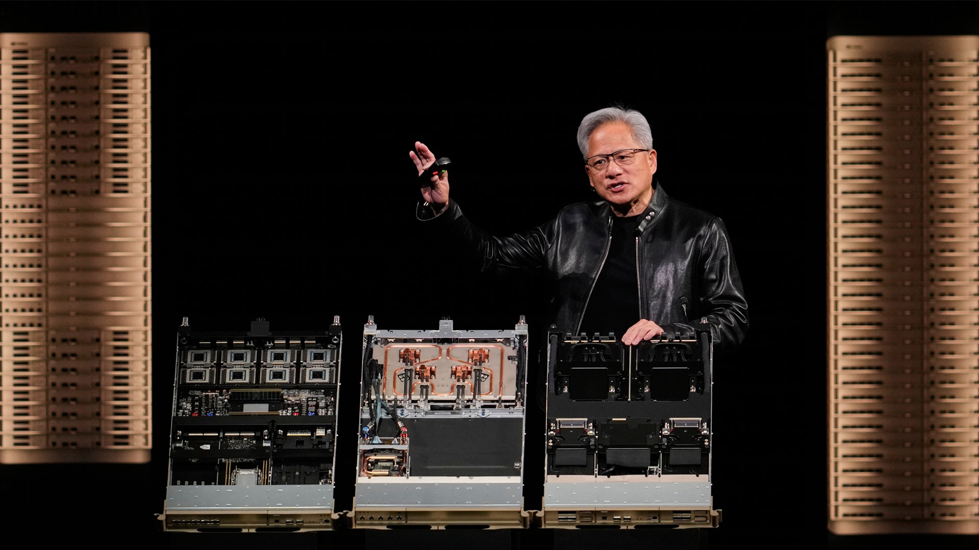 En man presenterar elektroniska komponenter på en scen. Han bär glasögon och svart läderrock, och pekar uppåt med ena handen. Bakom honom syns gyllene paneler och elektronisk utrustning.