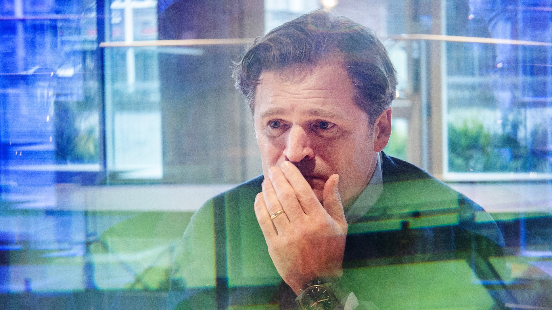 En man i grön skjorta står framför en modern glaskonstruktion och har handen vid munnen i en tankfull eller oroad pose. Bakgrunden visar blå och gröna reflektioner från byggnaden.