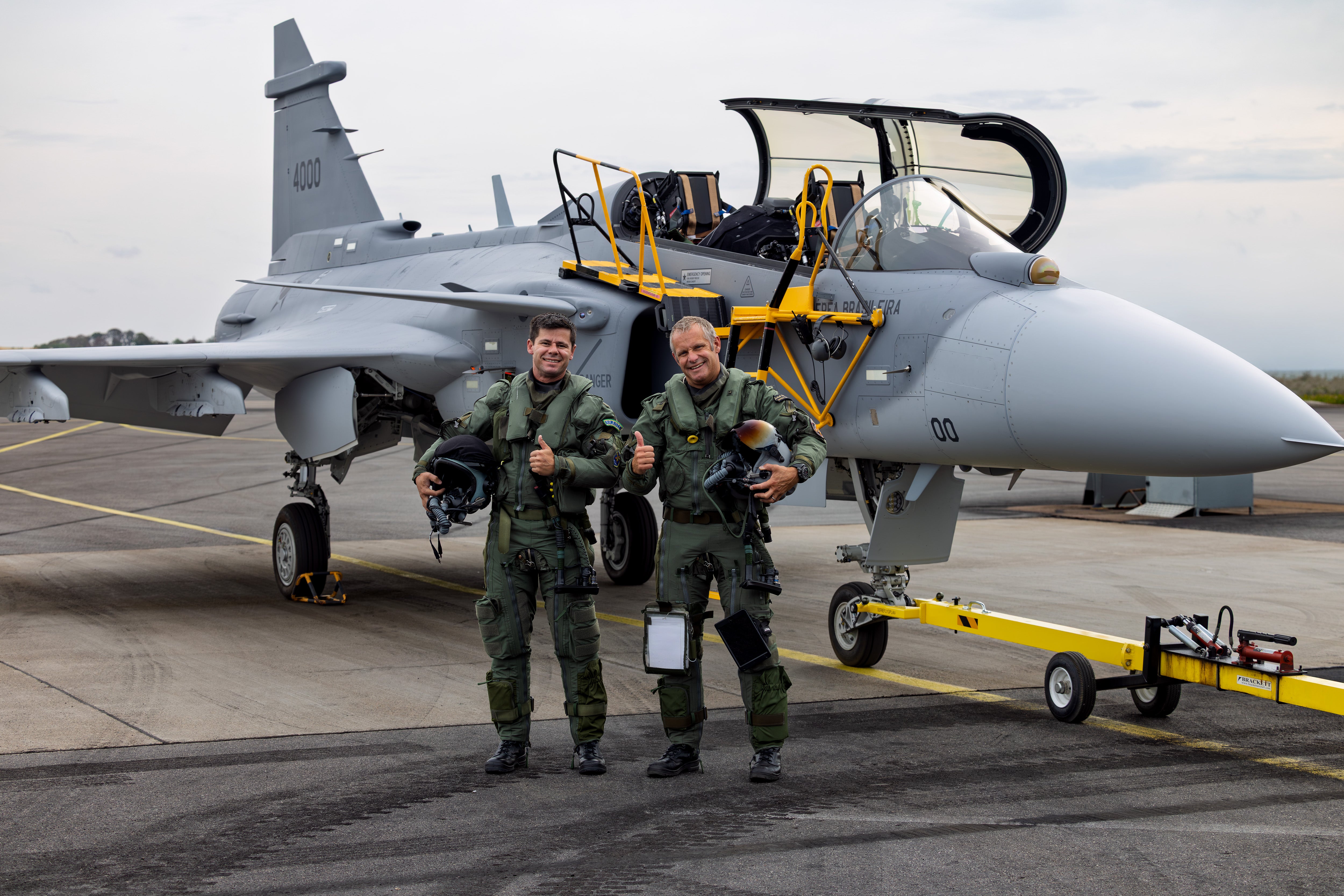 Två militärpiloter i gröna flightdräkter står framför ett grått stridsflygplan på en flygbas.