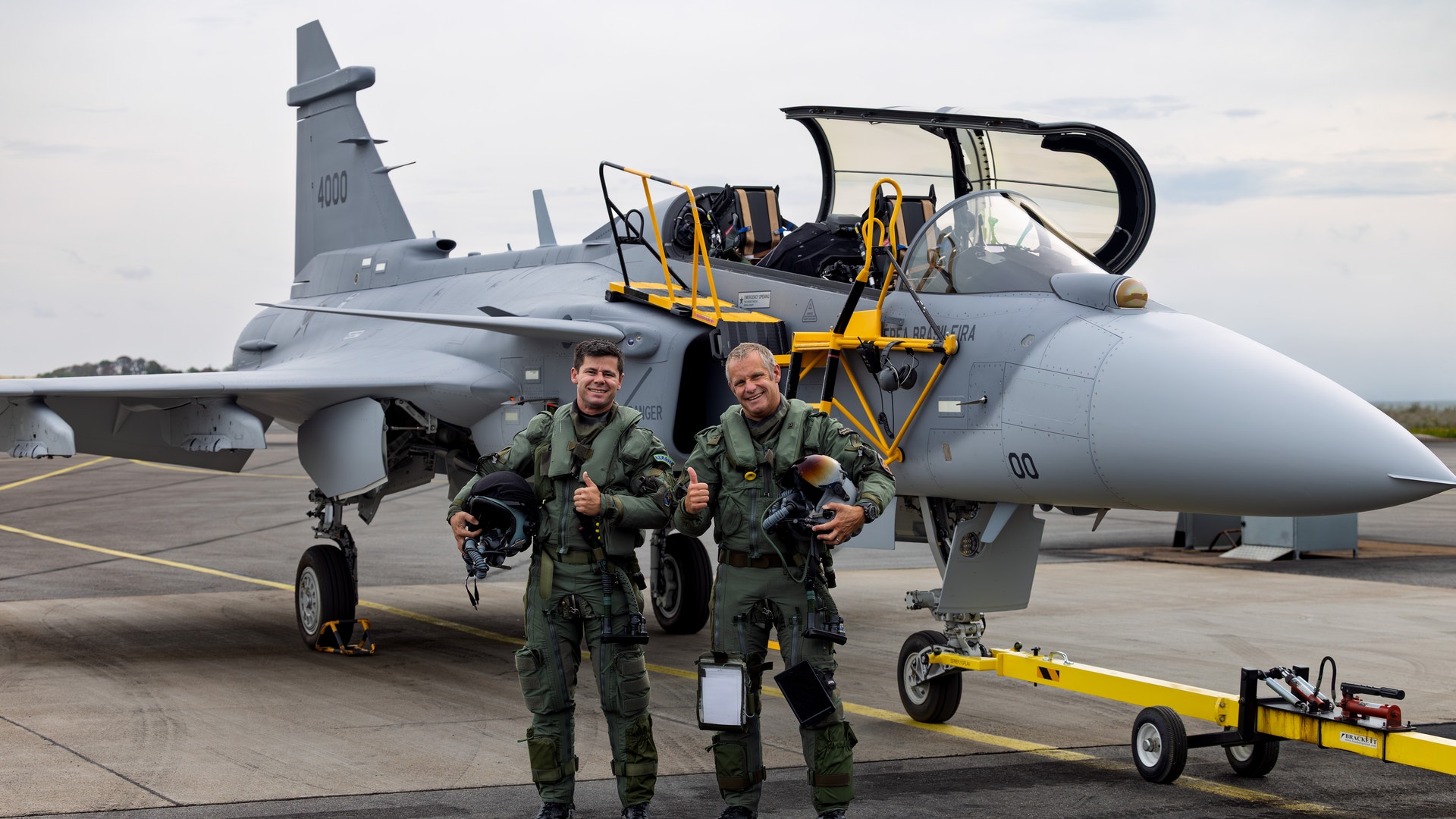 Två militärpiloter i gröna flightdräkter står framför ett grått stridsflygplan på en flygbas.