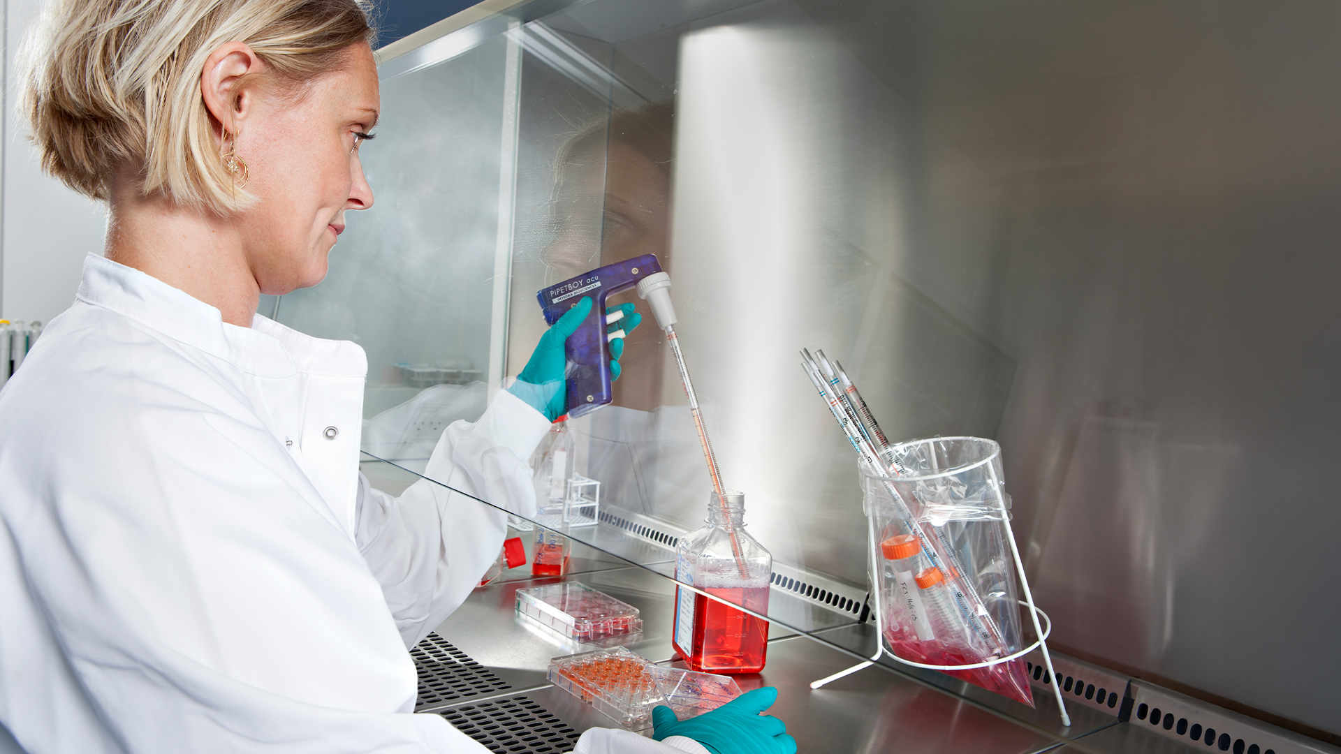 Laboratoriearbetare i vit rock arbetar i ett biosäkerhetsskåp med röda vätskeprover och laboratorieglasögon.