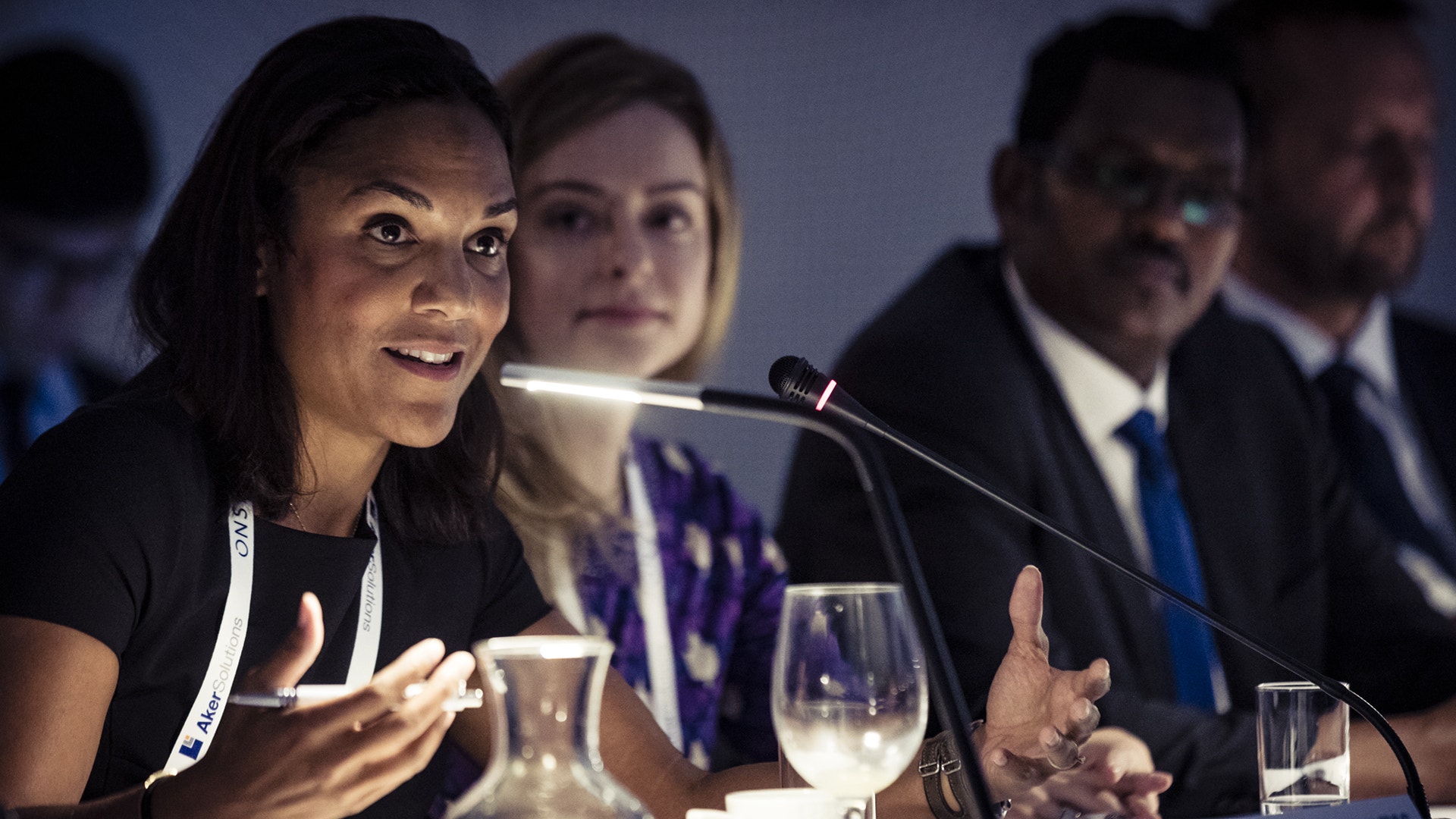 Kvinna talar vid en mikrofon under en konferens eller paneldiskussion, med andra deltagare sittande bakom henne vid ett bord.