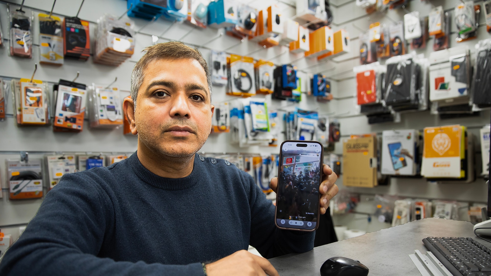 En man i mörkblå tröja står bakom en butiksdisk i en teknikbutik och håller upp en mobiltelefon. Bakom honom hänger mobiltillbehör på väggen.