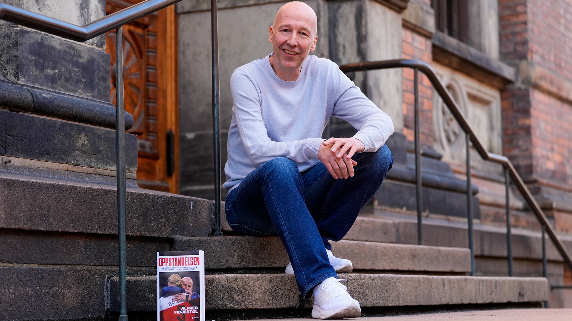 Porträtt av en man som sitter på trappsteg framför en tegelvägg. Han bär ljusblå tröja och mörkblå jeans, och ler mot kameran.
