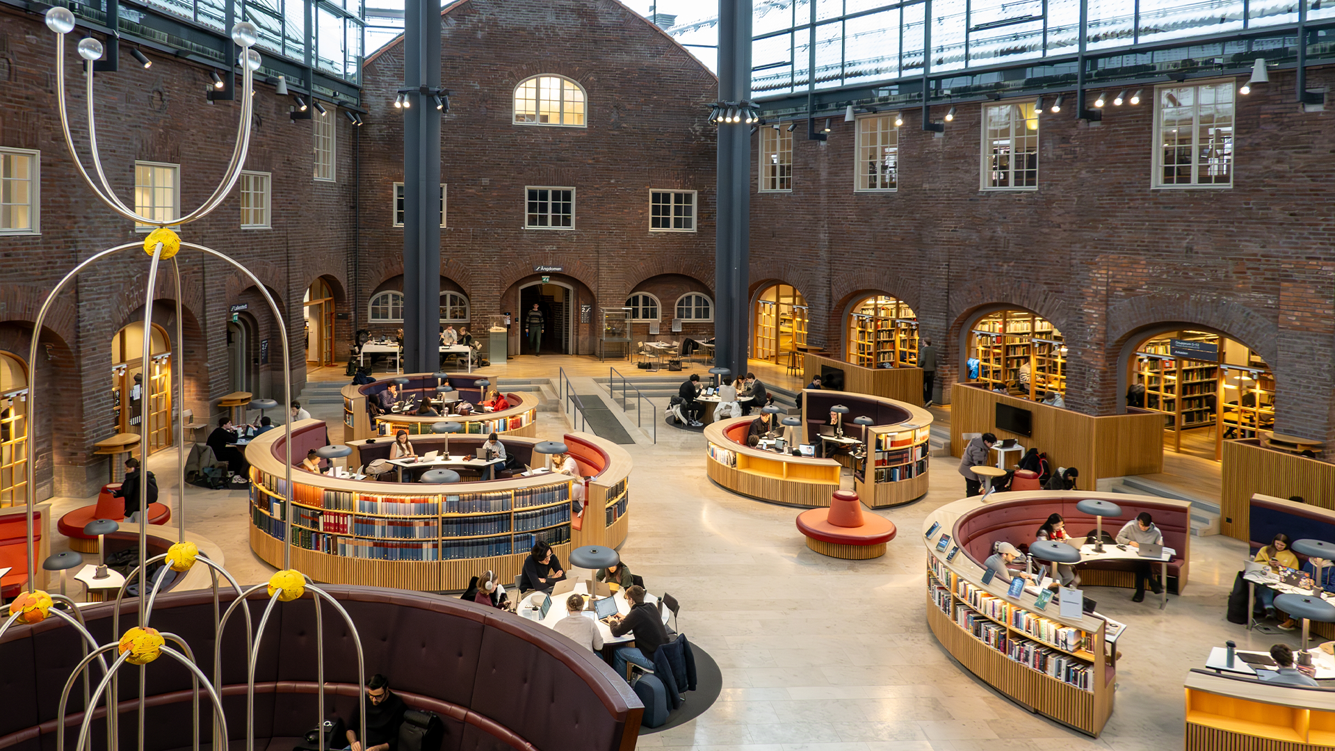 Modernt biblioteksinteriör med bökkällor organiserade i böjda trähyllor. Besökare läser och arbetar i det ljusa, öppna rummet omgivet av tegelvägg och arkitektonisk design.