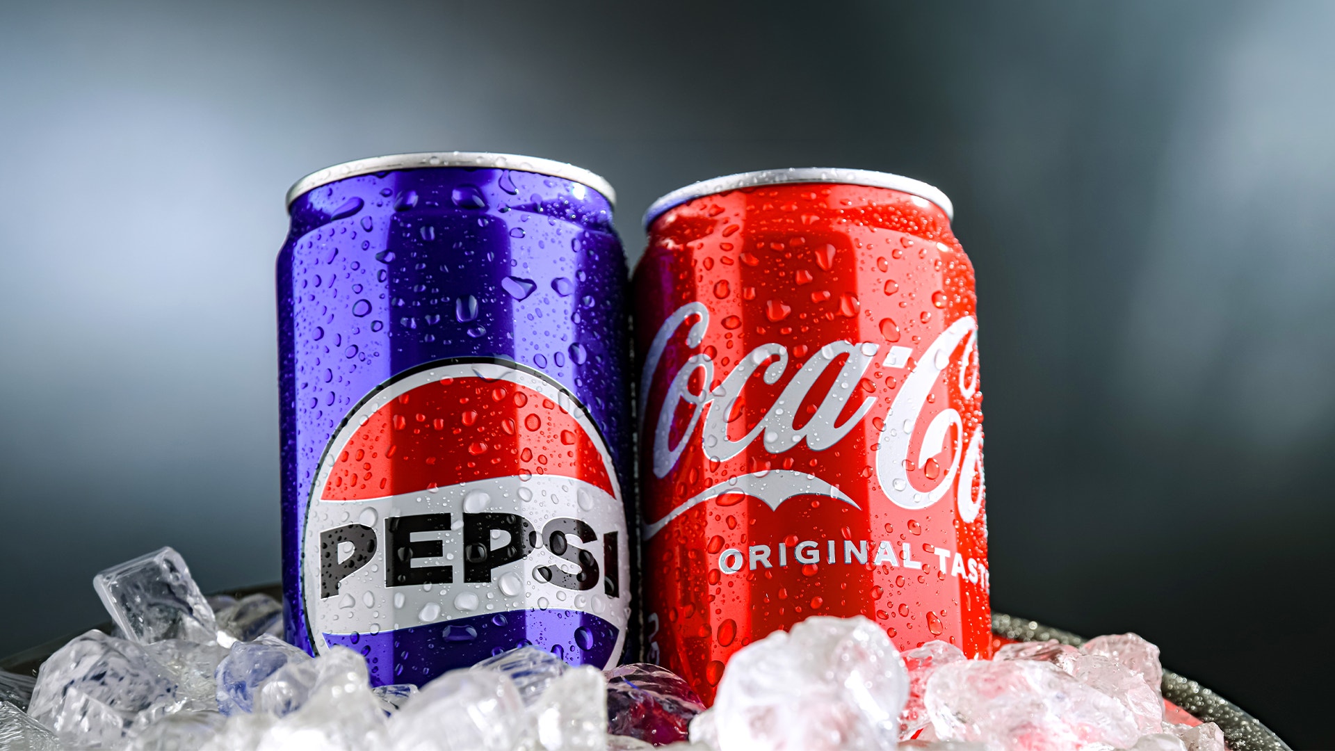 En blå Pepsi-burk och en röd Coca-Cola-burk står bredvid varandra bland isbitar. Burkarna är täckta av vattendroppar.