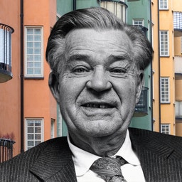 Svartvit porträttfoto av en äldre man i kostym och slips, monterad framför ett färgglatt urbant buskehus med orange, gula och gröna fasader.