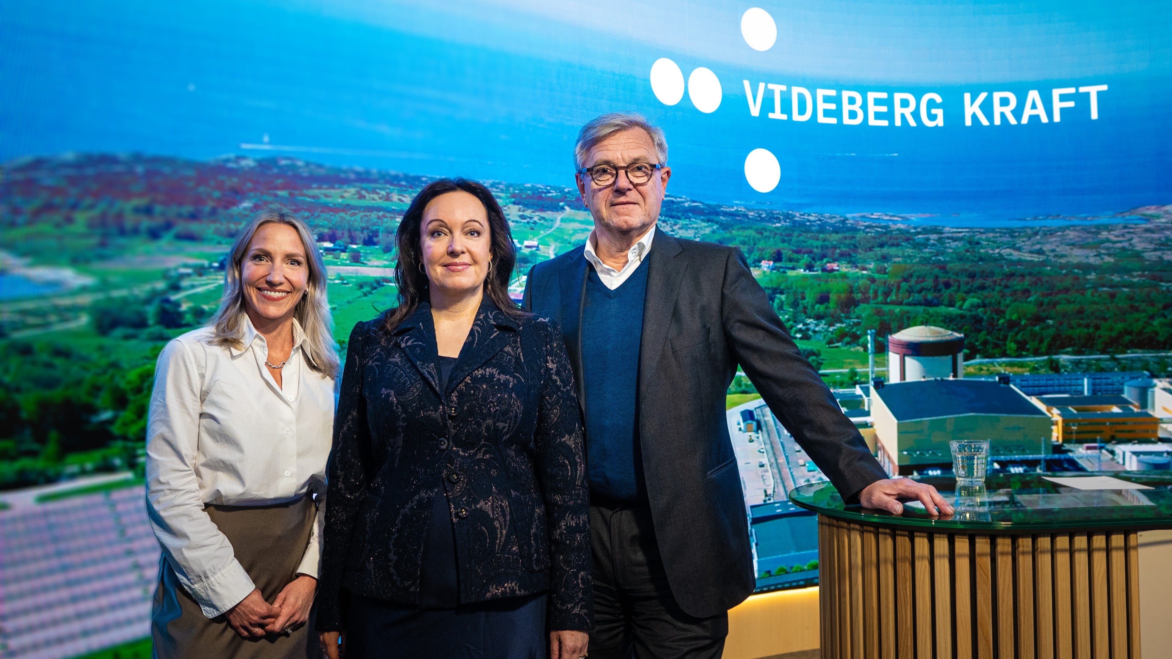 Tre personer står framför en bakgrund med Videberg Kraft-logotyp och ett landskap med kraftverk.