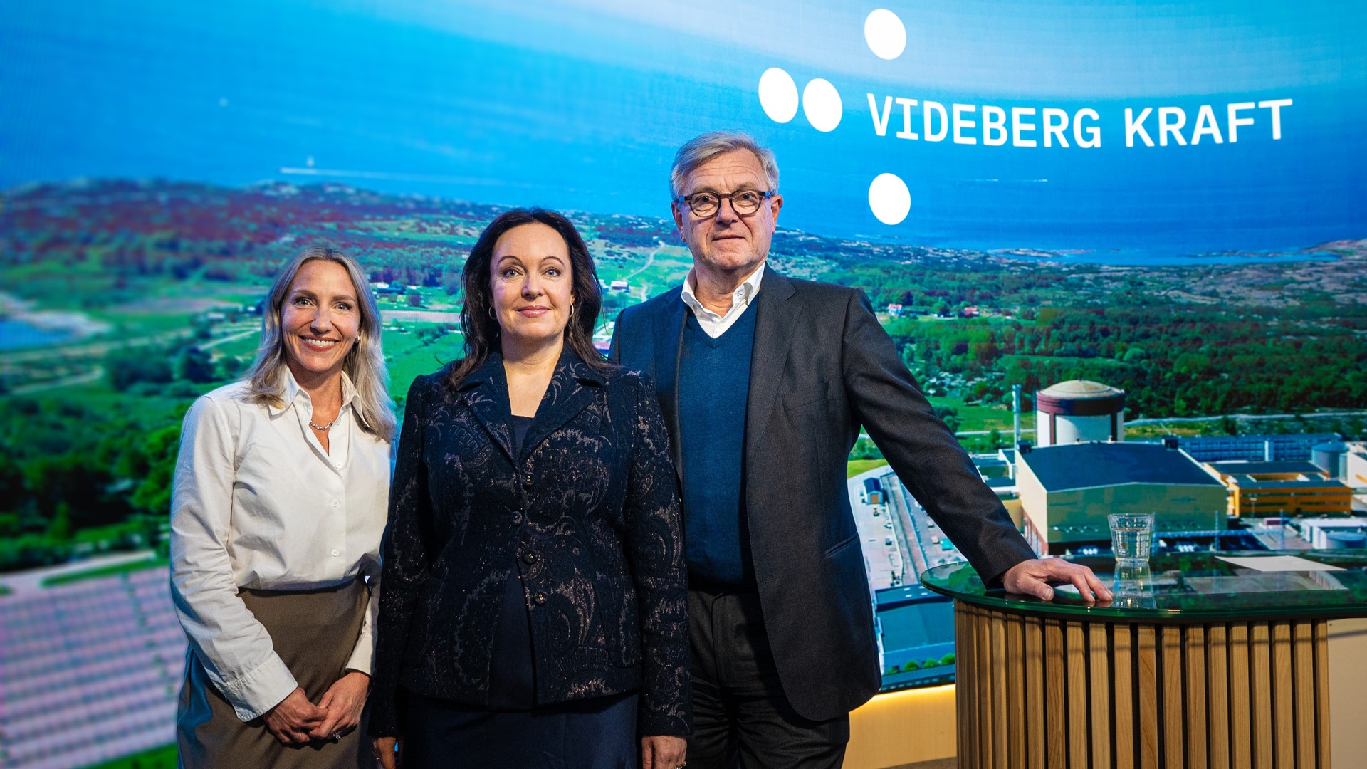 Tre personer står framför en bakgrund med Videberg Kraft-logotyp och ett landskap med kraftverk.
