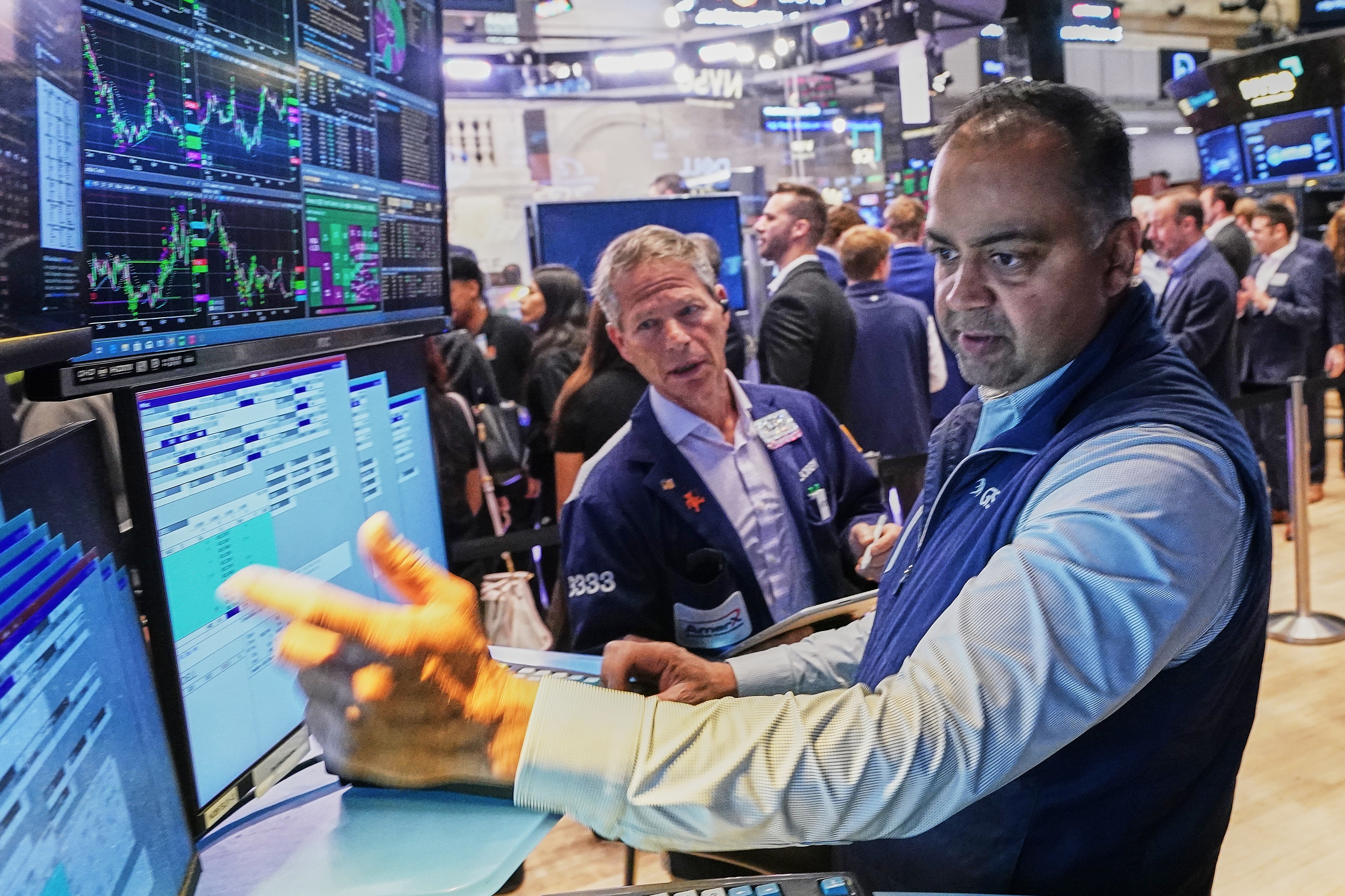 Män i affärskläder arbetar vid dataskärmar på en börs. Skärmarna visar finansiella grafer och diagram i blått och grönt. Flera andra personer syns i bakgrunden.