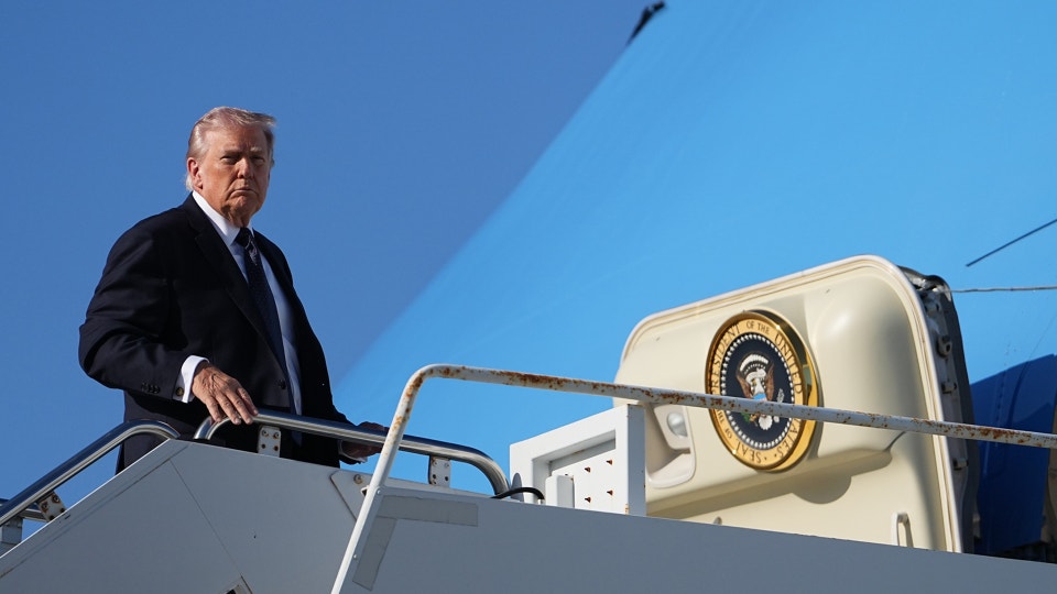 Man i mörk kostym på flygplanstrappan framför ett stort vitt flygplan mot blå himmel.