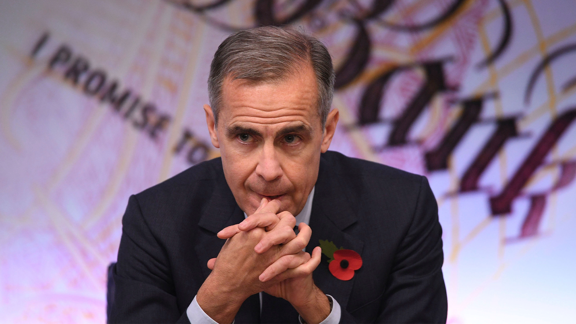 En man i mörk kostym med röd vallmoblomma sitter och lyssnar uppmärksamt under en offentlig event.