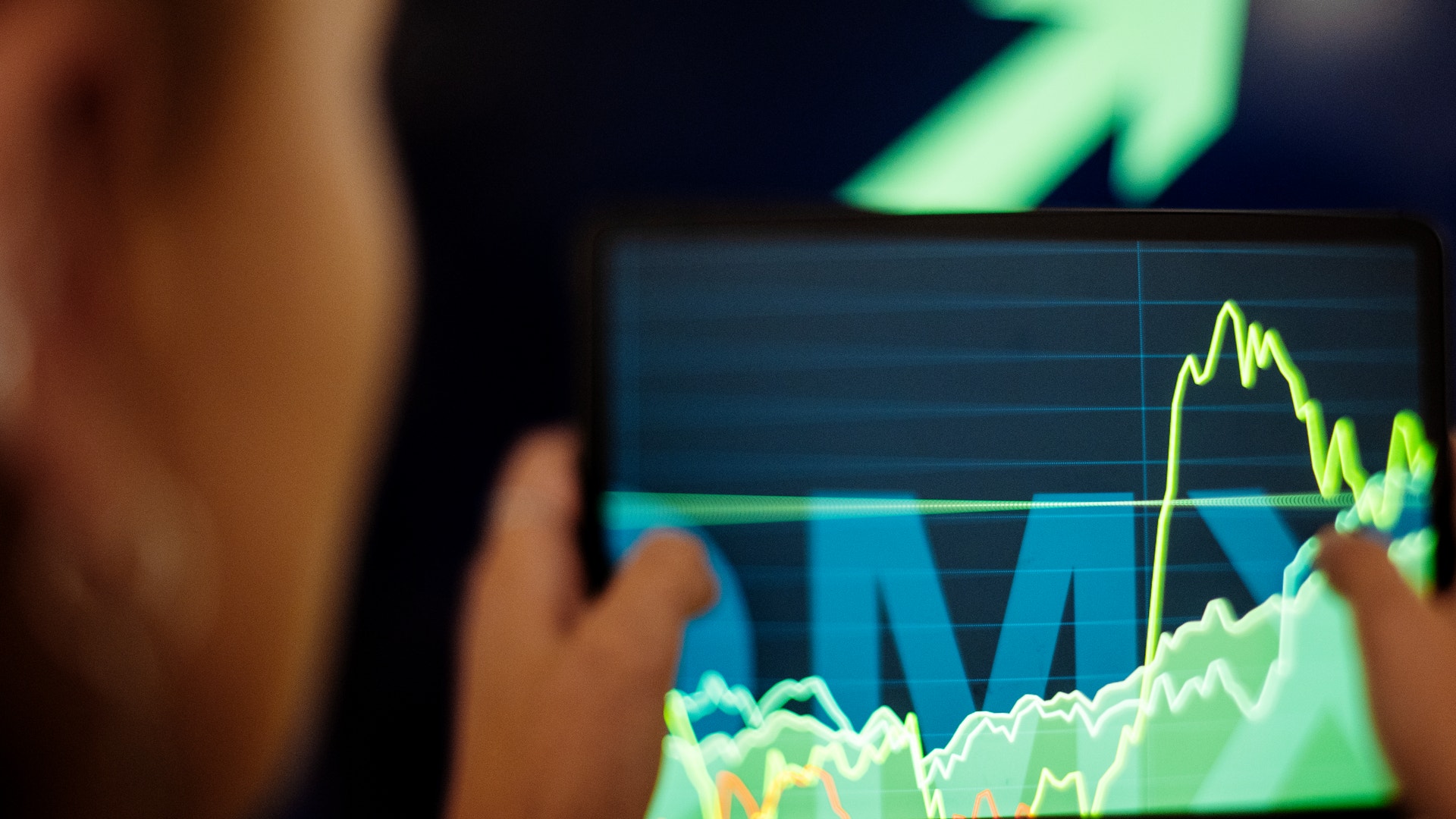 Person tittar på en datorskärm med finansiella grafer och diagram som visar aktiekurser och marknadstrender i grönt och gult.