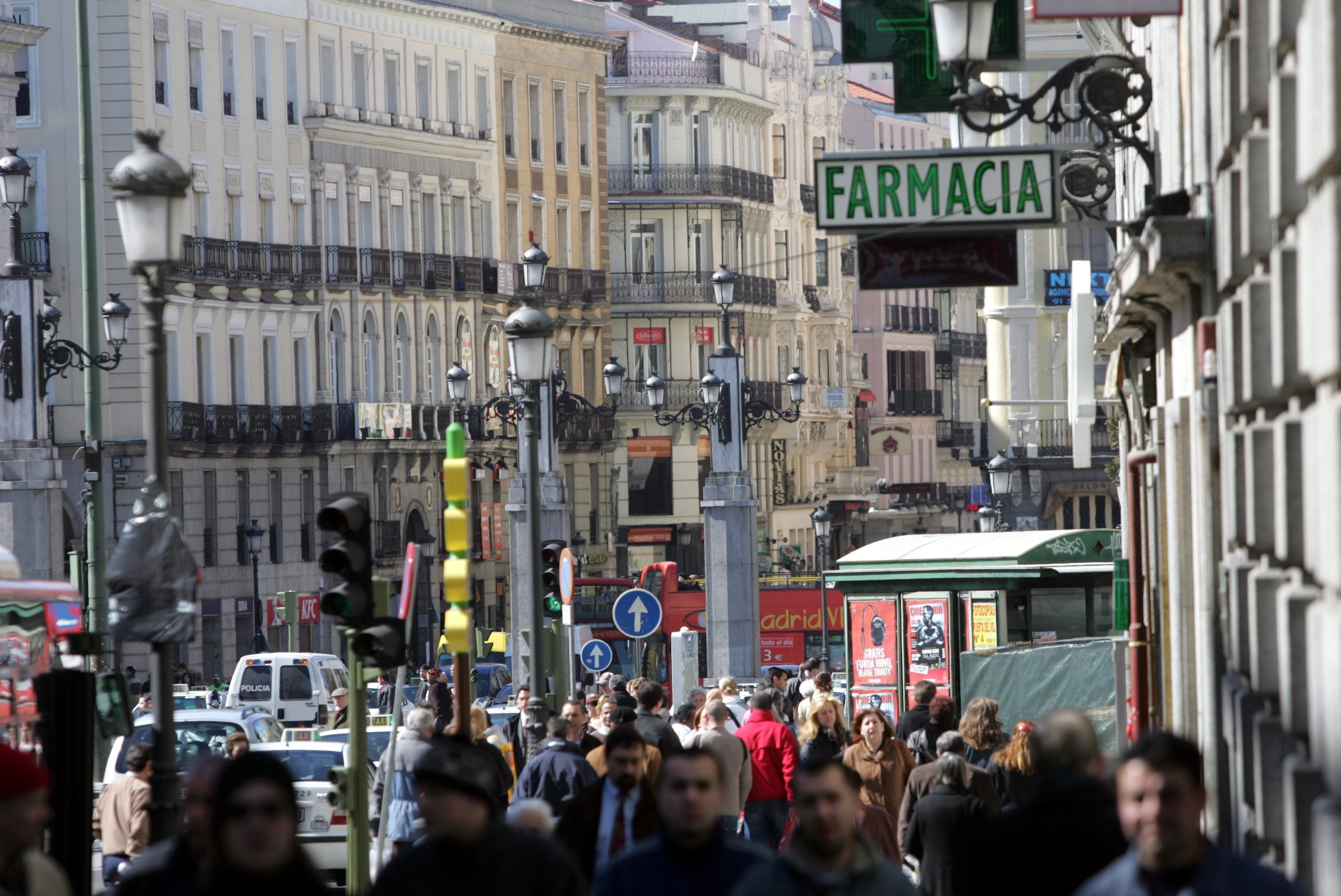Folkmassa på en livlig gata i en stad, med äldre byggnader och en skylt där det står Farmacia.