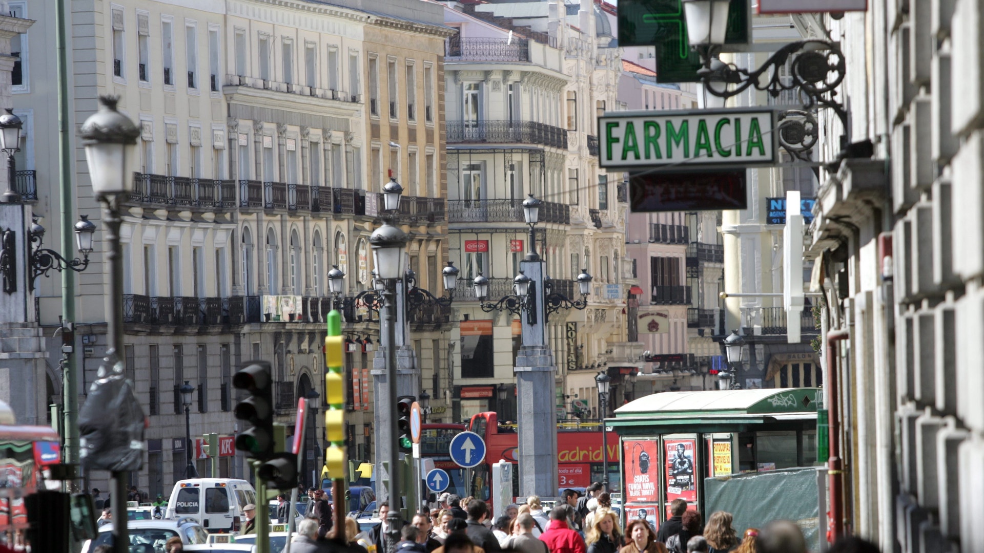 Folkmassa på en livlig gata i en stad, med äldre byggnader och en skylt där det står Farmacia.