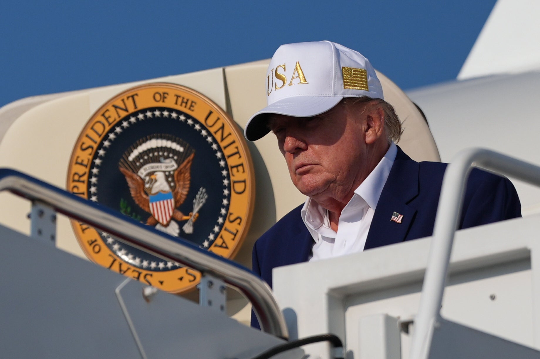 Man i vit keps och mörk kostym står vid en flygplanstrappa framför ett flygplan med presidentens sigill.
