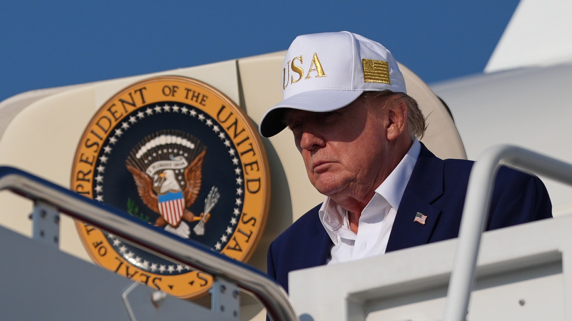 Man i vit keps och mörk kostym står vid en flygplanstrappa framför ett flygplan med presidentens sigill.
