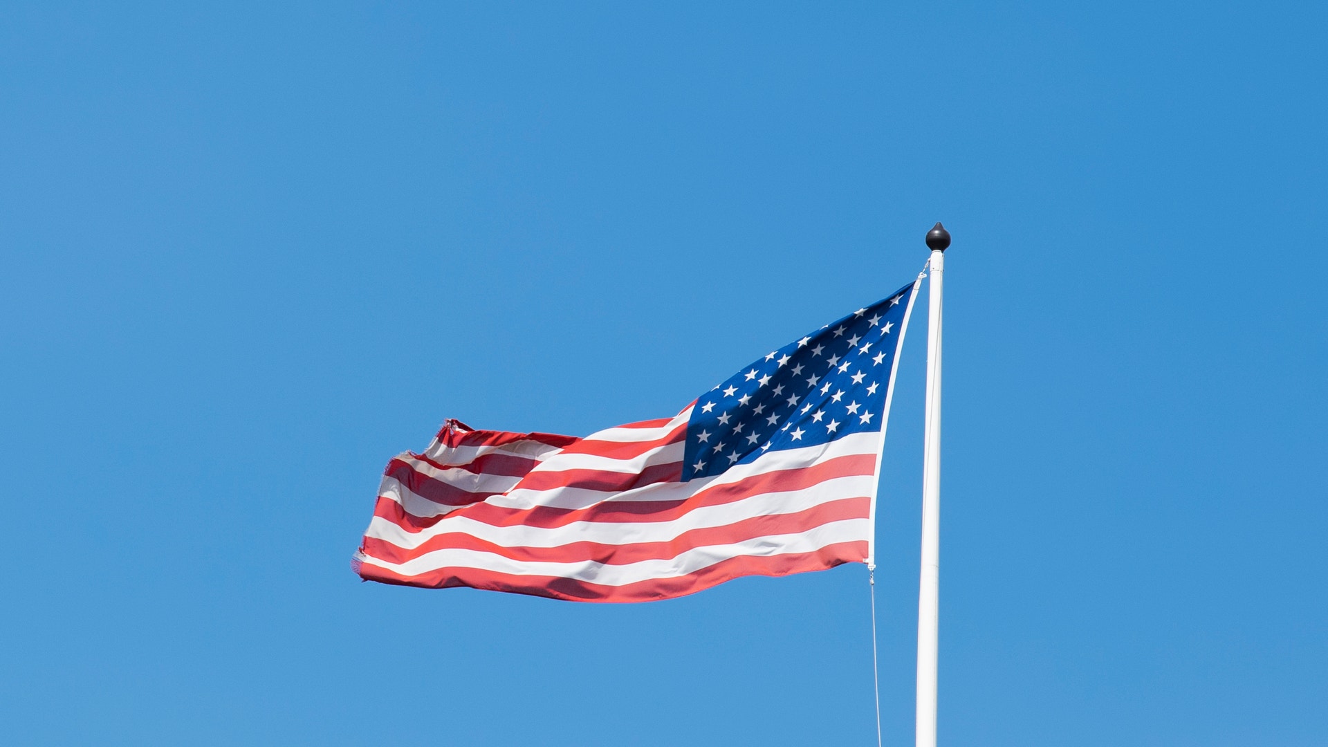 Amerikansk flagga som vajar på en vit flaggstång mot en klar blå himmel.