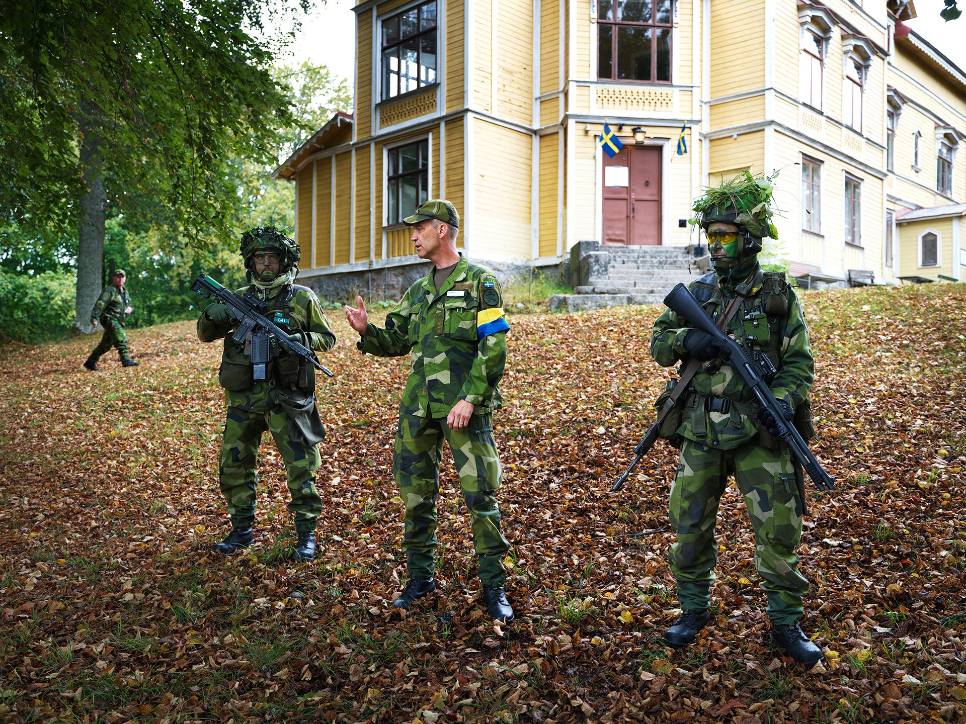 Soldater i kamouflageuniform och beväpnade med gevär står utanför en gul träbyggnad, omgivna av höstlöv på marken.