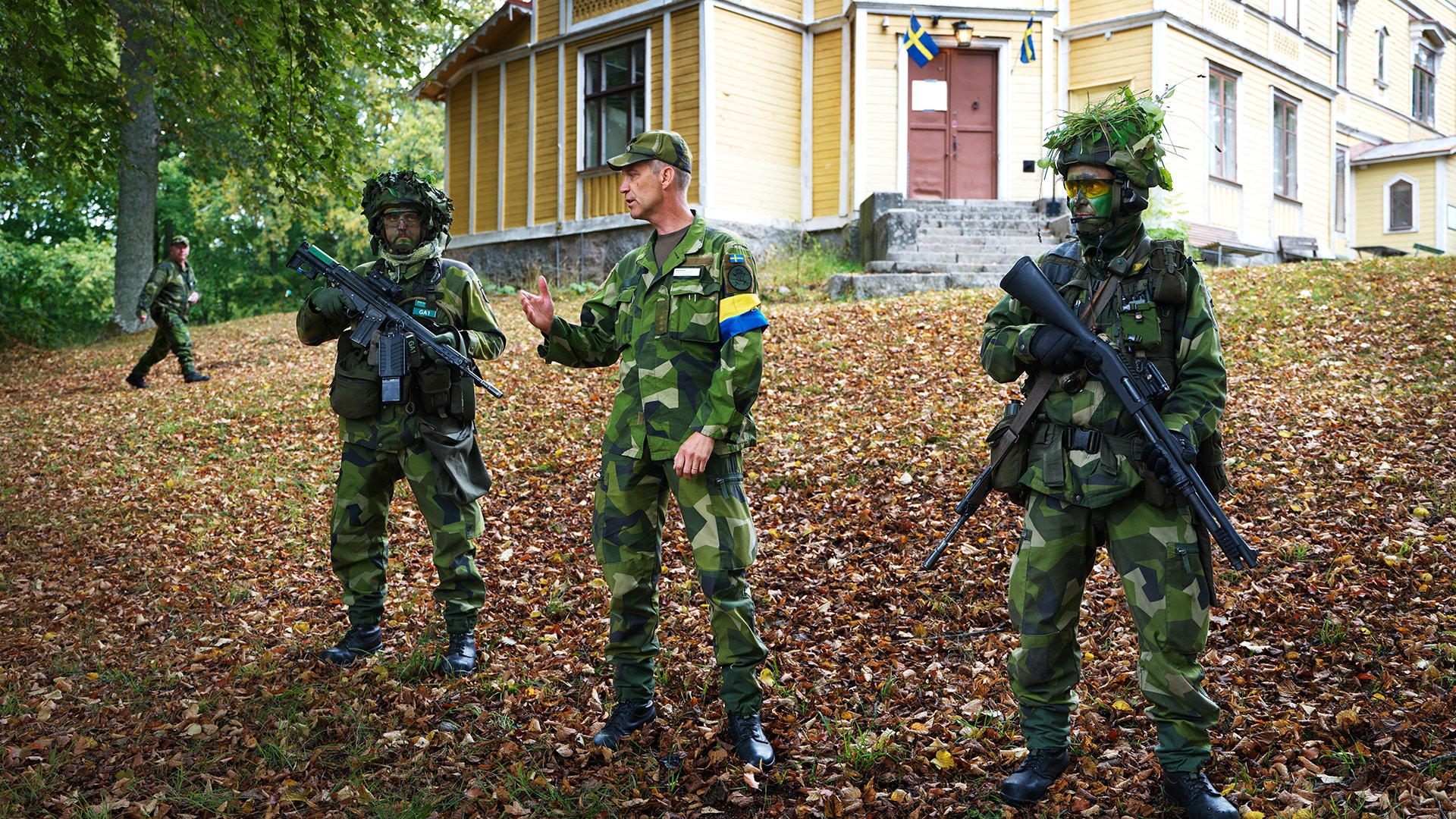 Soldater i kamouflageuniform och beväpnade med gevär står utanför en gul träbyggnad, omgivna av höstlöv på marken.