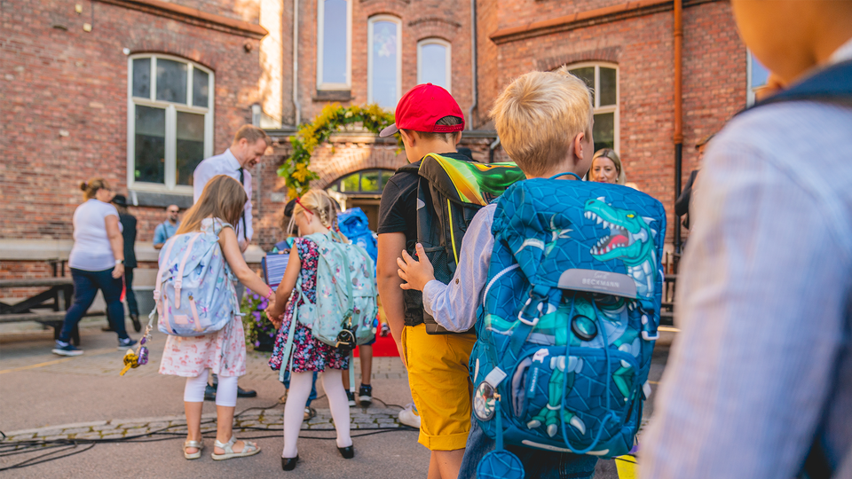 Fem barn går mot skolentrén för att börja skolan igen efter sommarlovet. Barnen har alla olika färgglada ryggsäckar och går mot en en lärare som hälsar dem välkomna.