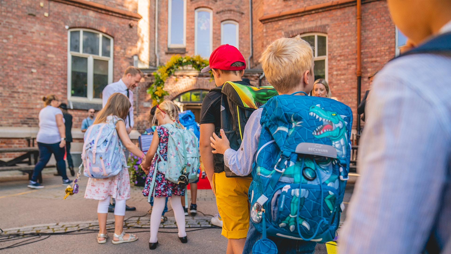 Fem barn går mot skolentrén för att börja skolan igen efter sommarlovet. Barnen har alla olika färgglada ryggsäckar och går mot en en lärare som hälsar dem välkomna.