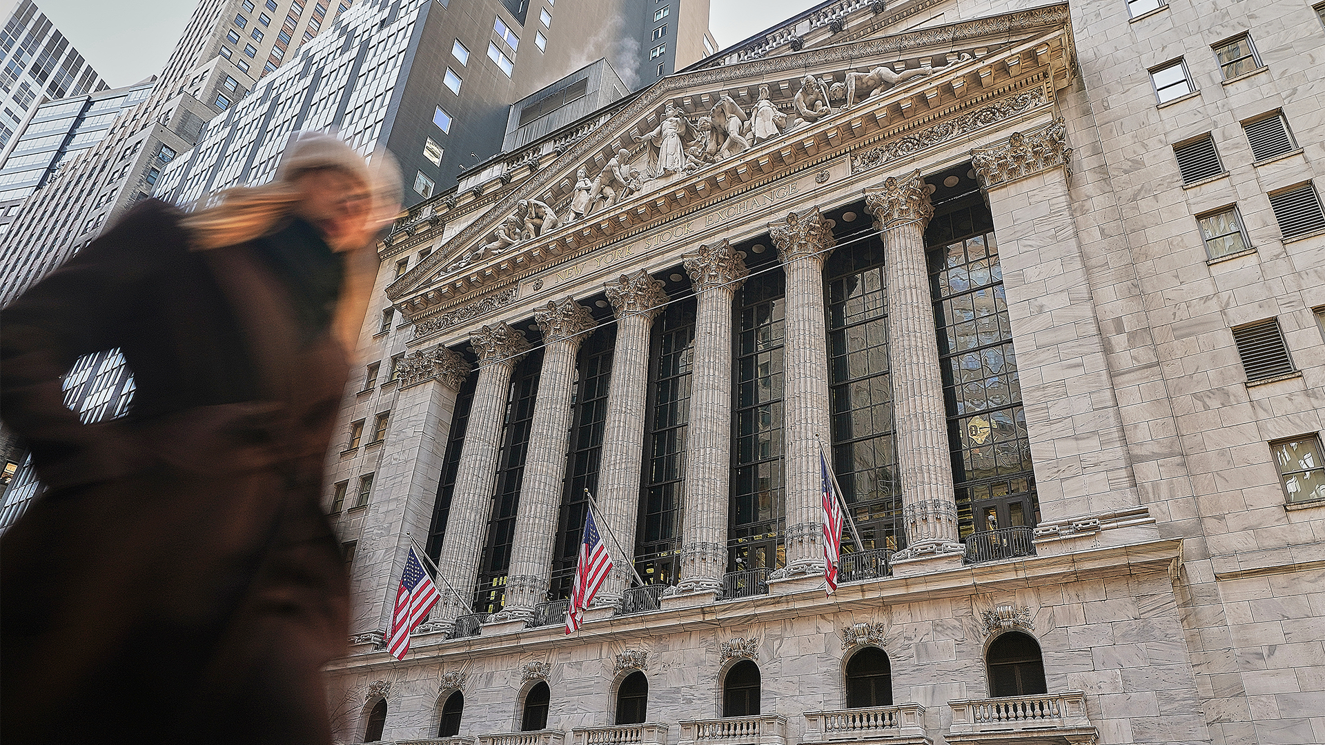 En person i förgrunden framför New York Stock Exchange-byggnaden, en imponerande neoklassisk arkitektur med höga vita kolonner, ornamenterad fasad och amerikanska flaggor som hänger på byggnaden. Moderna skyskrapor syns i bakgrunden.