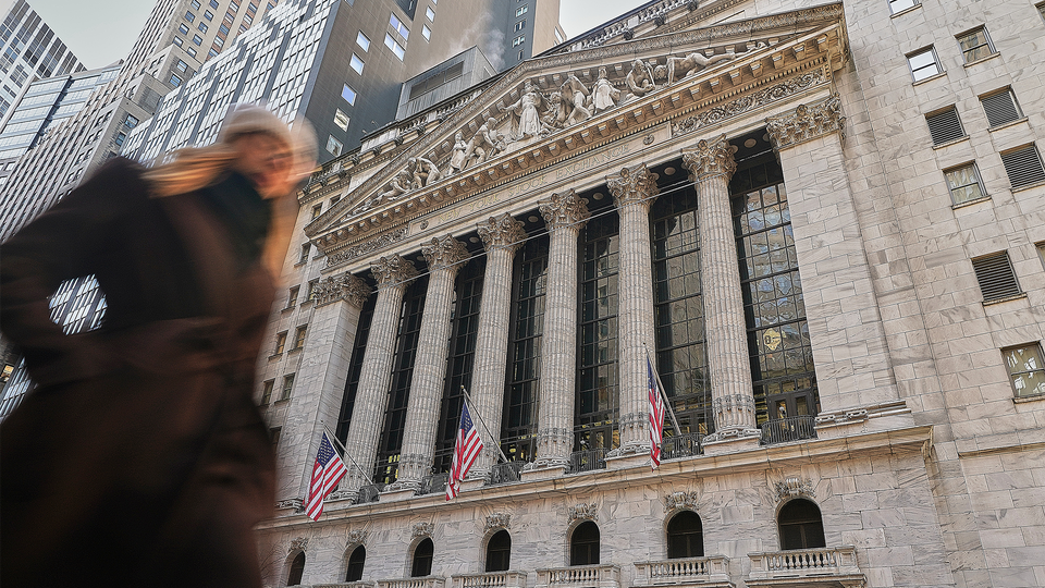 En person i förgrunden framför New York Stock Exchange-byggnaden, en imponerande neoklassisk arkitektur med höga vita kolonner, ornamenterad fasad och amerikanska flaggor som hänger på byggnaden. Moderna skyskrapor syns i bakgrunden.