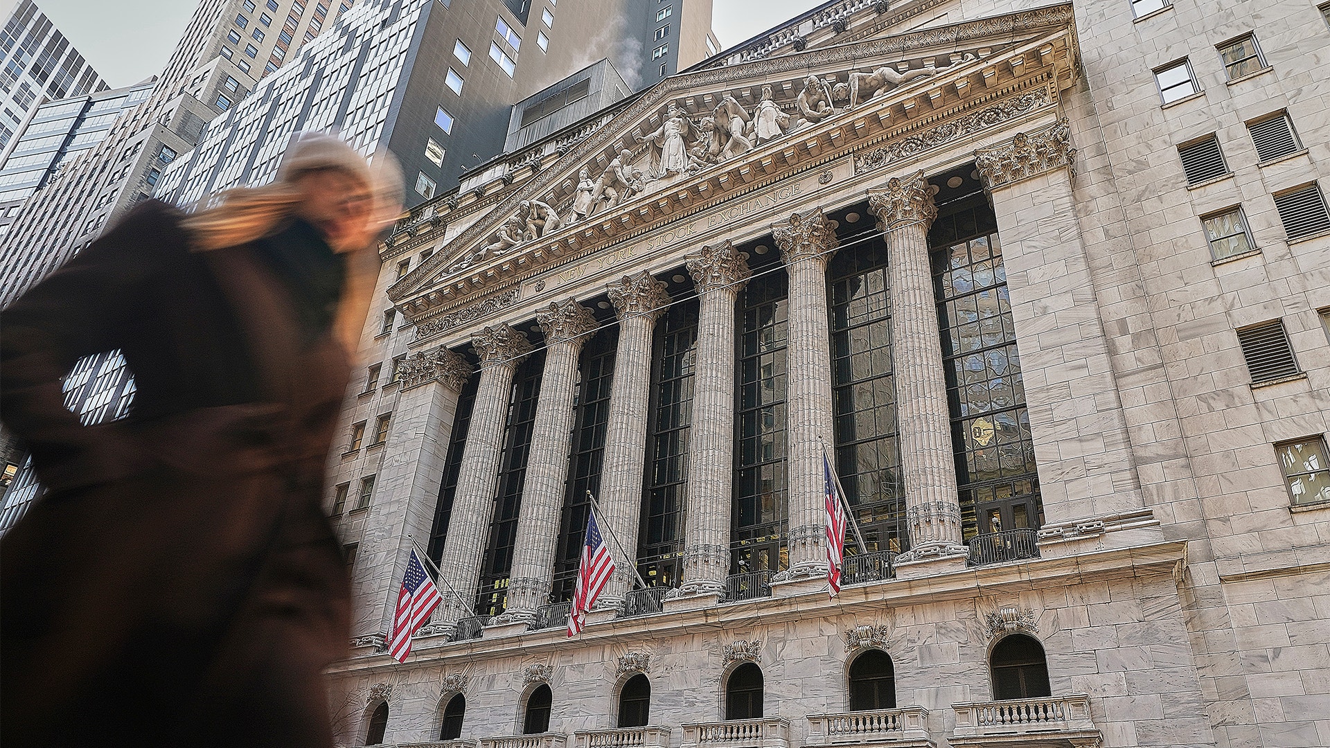 En person i förgrunden framför New York Stock Exchange-byggnaden, en imponerande neoklassisk arkitektur med höga vita kolonner, ornamenterad fasad och amerikanska flaggor som hänger på byggnaden. Moderna skyskrapor syns i bakgrunden.