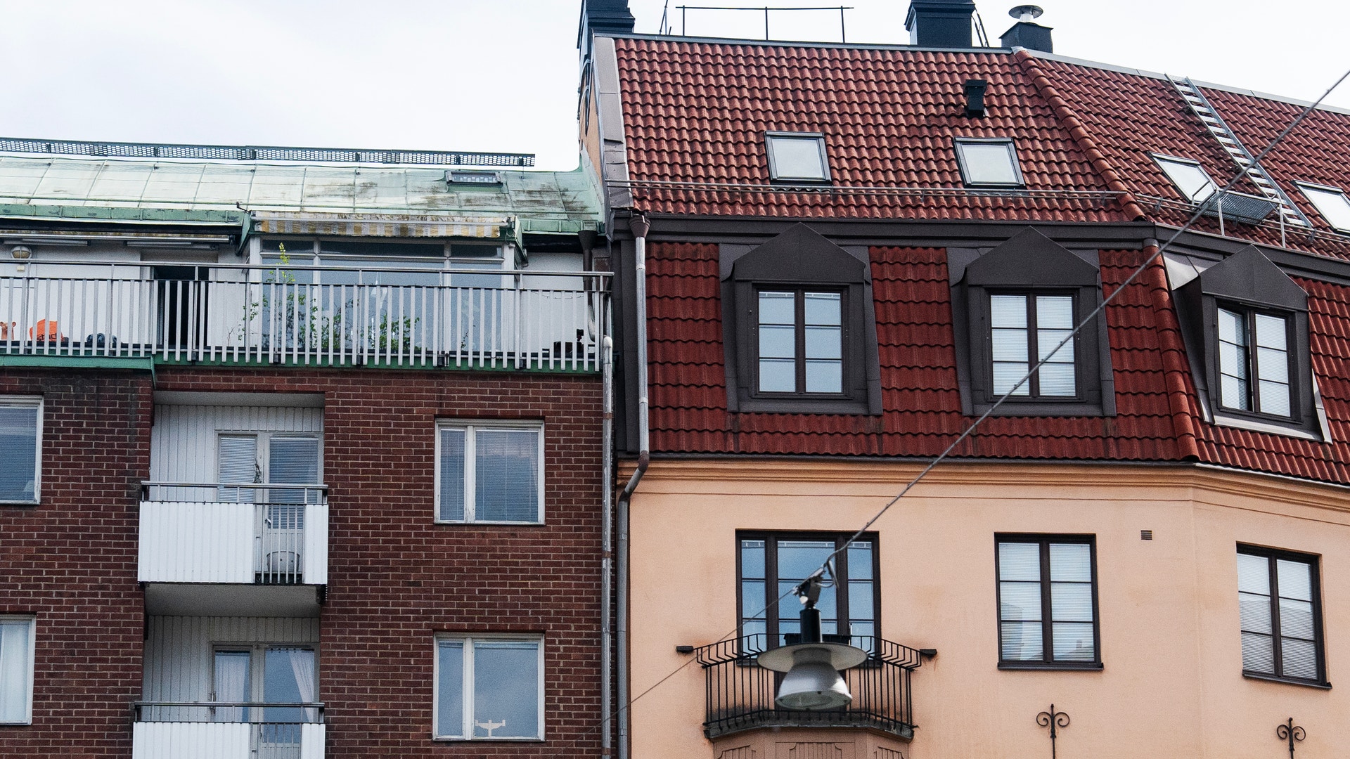 Två intilliggande bostadshus med olika fasader. Till vänster ett rödbrunt tegelshus med vita balkonger, till höger ett gult hus med rött tegeltaket och svarta fönster.