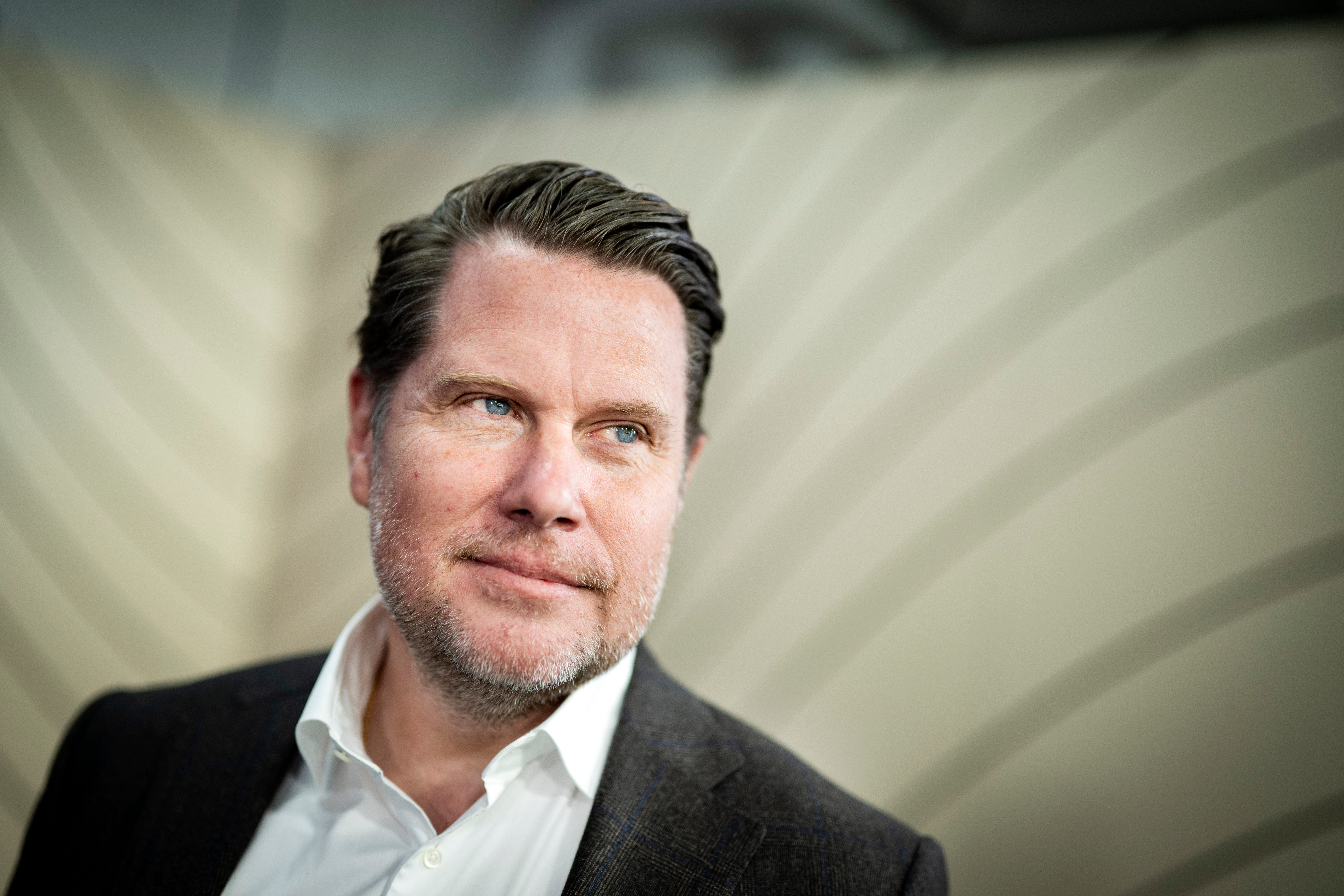 Porträttfoto av en man i mörk kavaj och vit skjorta mot en neutral bakgrund.