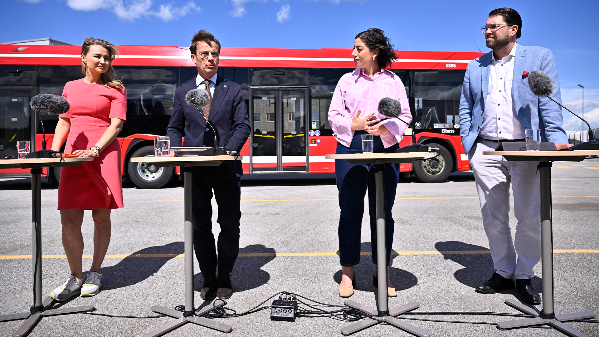 Fyra personer vid mikrofoner framför en röd buss under en presskonferens.