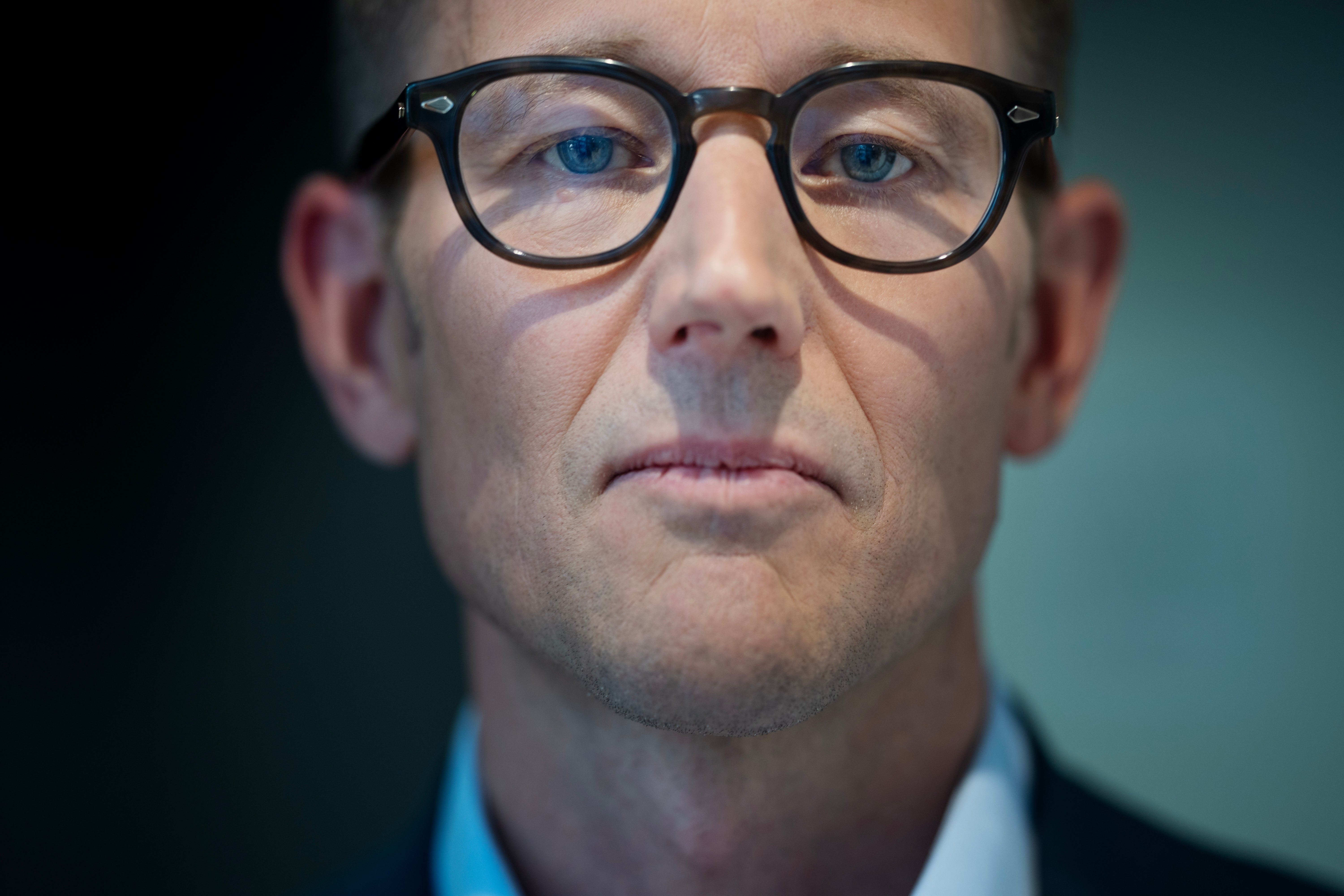 Porträtt av en man med glasögon och blå ögon som tittar direkt in i kameran. Han bär en blå skjorta mot en suddig grönaktig bakgrund.