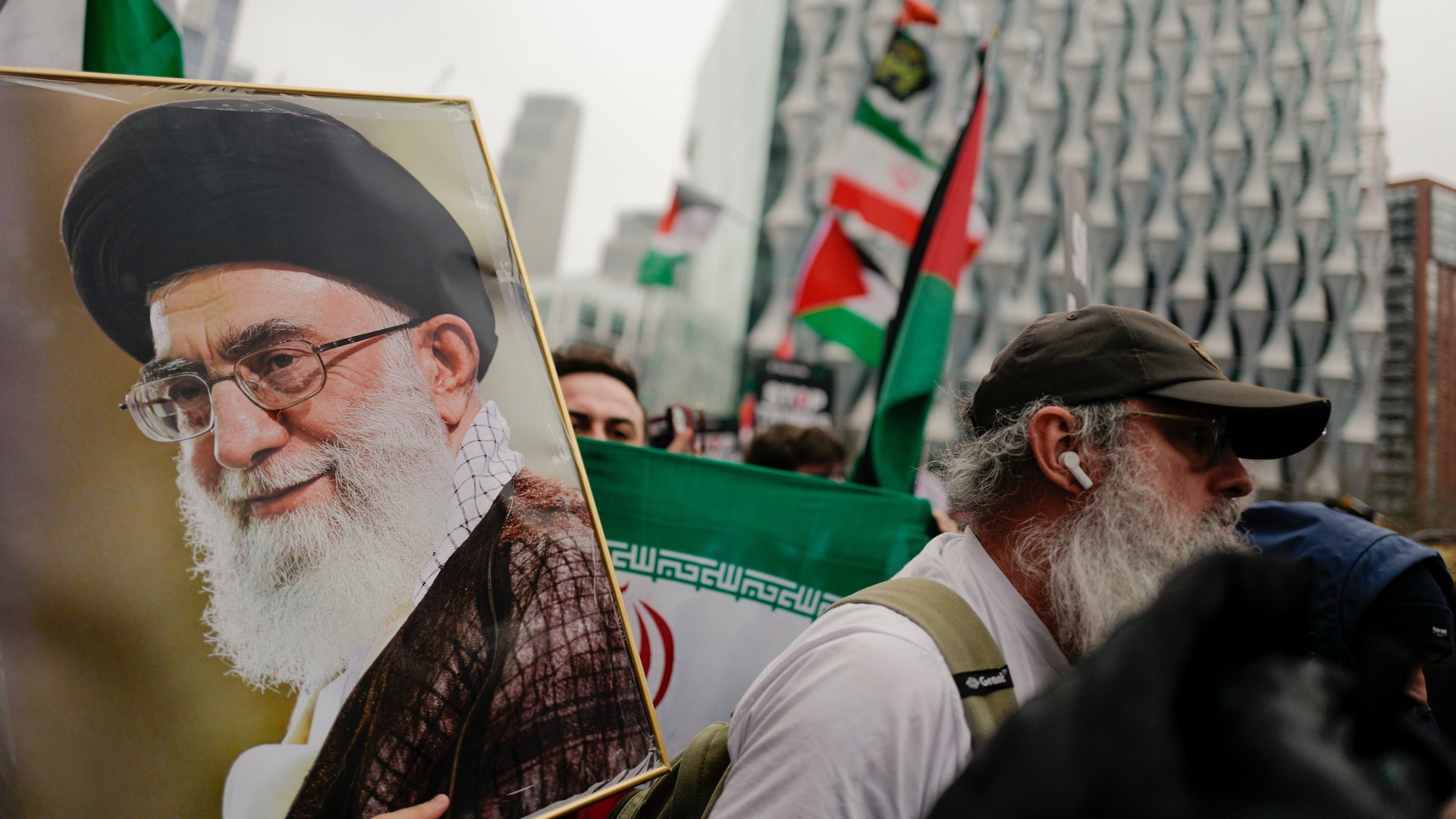 En offentlig samling med iranska flaggor och en porträttbild av en man med skägg och glasögon.