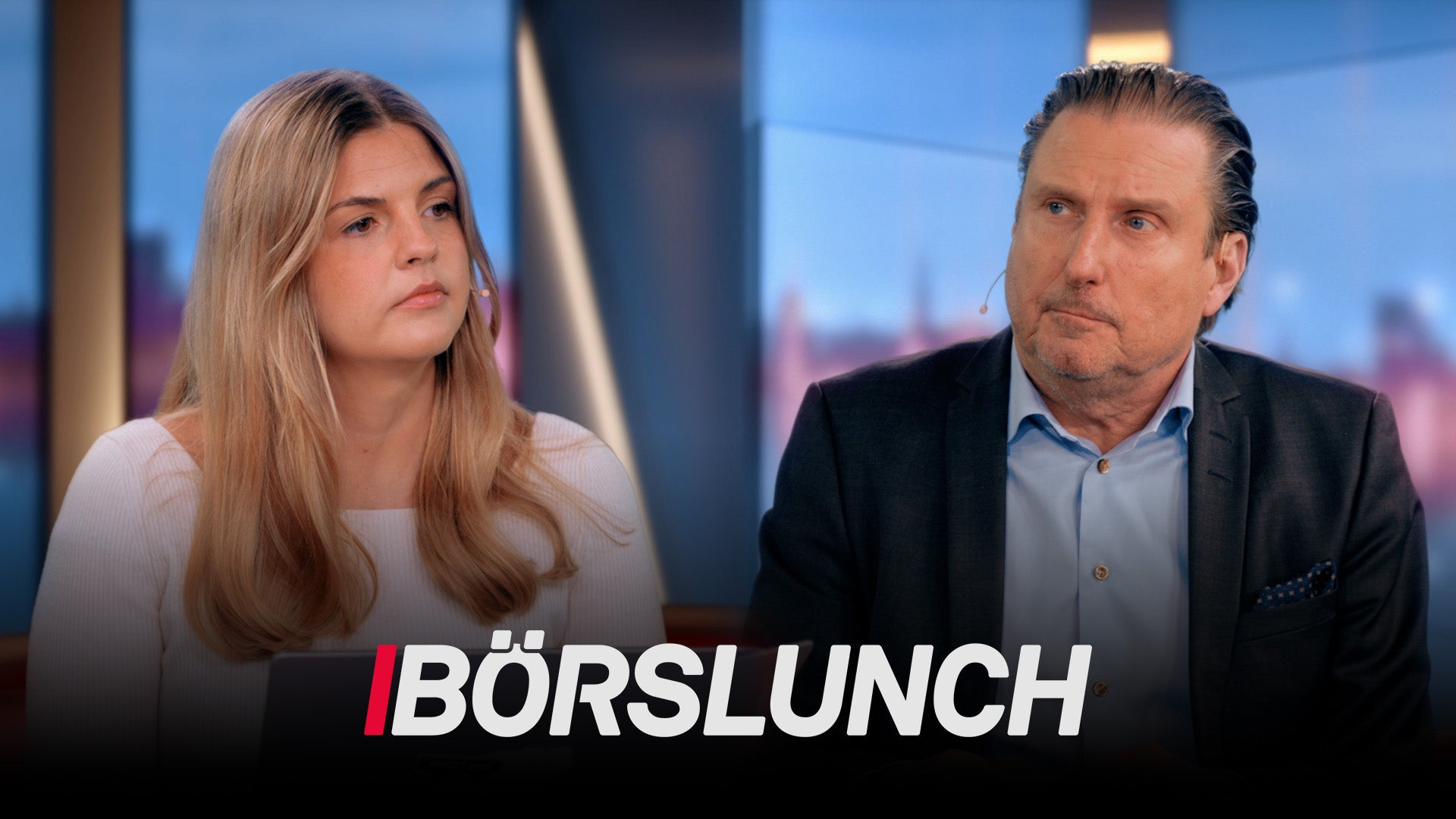 En kvinna och en man sitter vid ett studiobord under inspelning av TV-programmet Börslunch. Kvinnan till vänster har långt ljusbrunt hår och bär en ljus tröja. Mannen till höger är klädd i mörk kavaj och ljusblå skjorta. I bakgrunden syns en suddig stadshorisont. Programtiteln "BÖRSLUNCH" visas längst ned på bilden.