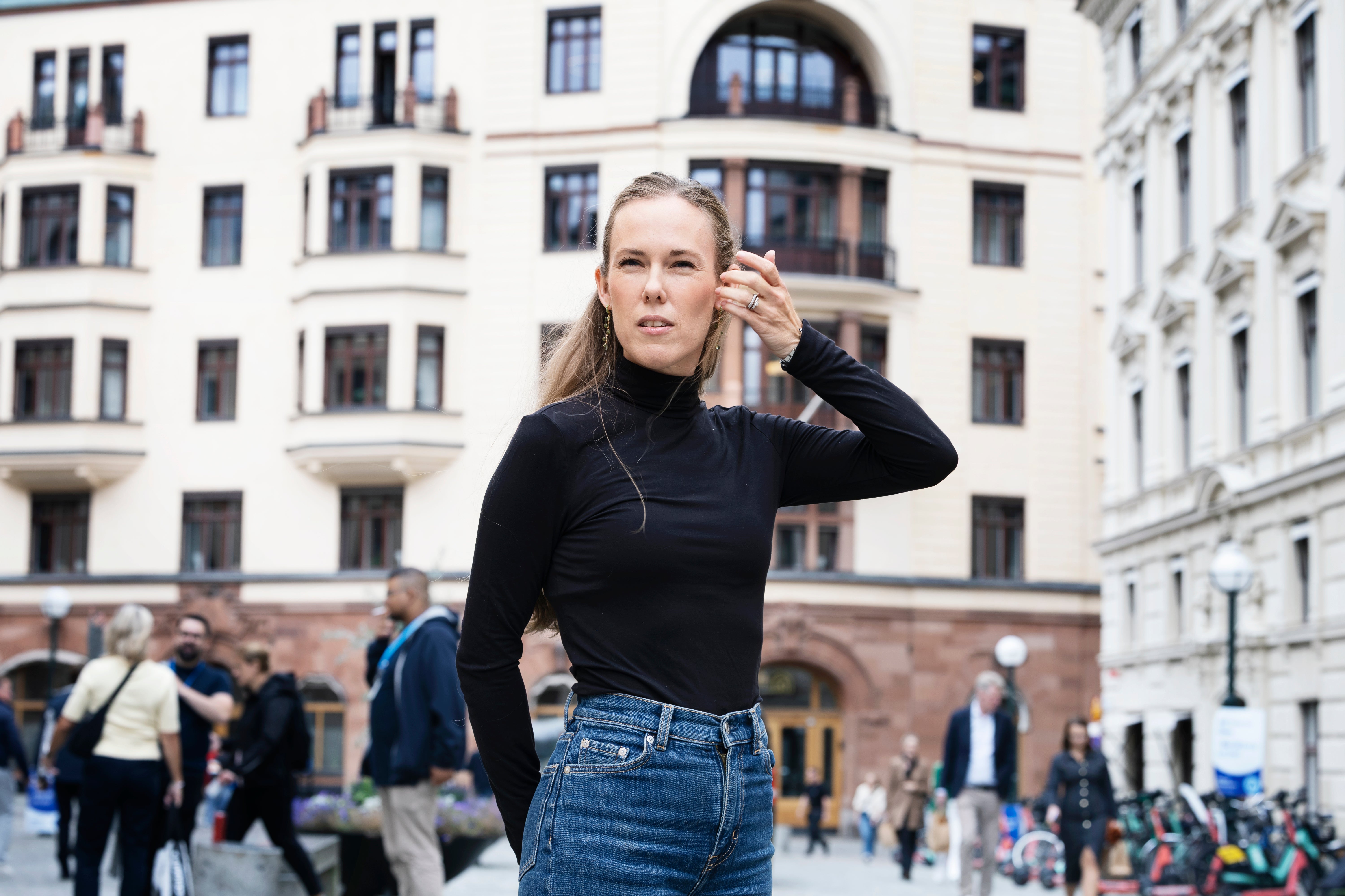Porträtt av en kvinna som står på en stadsplats. Hon bär svart långärmad tröja och jeans, och har handen vid sitt öra. Klassiska byggnadsfasader syns i bakgrunden tillsammans med andra människor på platsen.