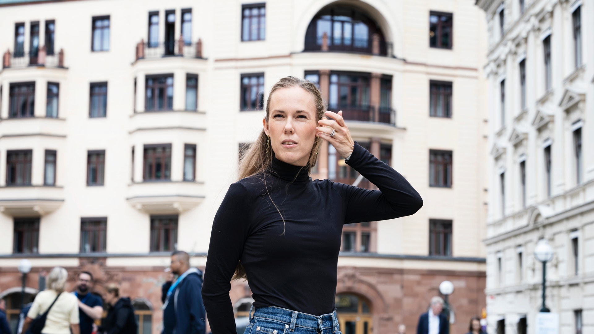 Porträtt av en kvinna som står på en stadsplats. Hon bär svart långärmad tröja och jeans, och har handen vid sitt öra. Klassiska byggnadsfasader syns i bakgrunden tillsammans med andra människor på platsen.