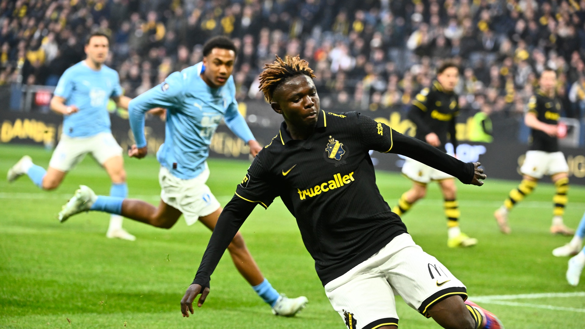 Zadok Yohanna i svart och gult dräkt springer med bollen under en match, medan motspelare i ljusblå försöker försvara. Publik syns i bakgrunden.