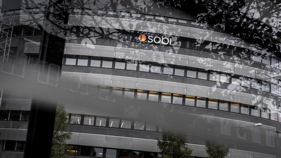 Modernt kontorsbyggnad med SOBI-logotyp på fasaden. Byggnaden har horisontella fönsterband och metallisk-glasarkitektur. Gröna växter vid byggnadens bas. Fotot är taget från låg vinkel uppåt mot himlen.