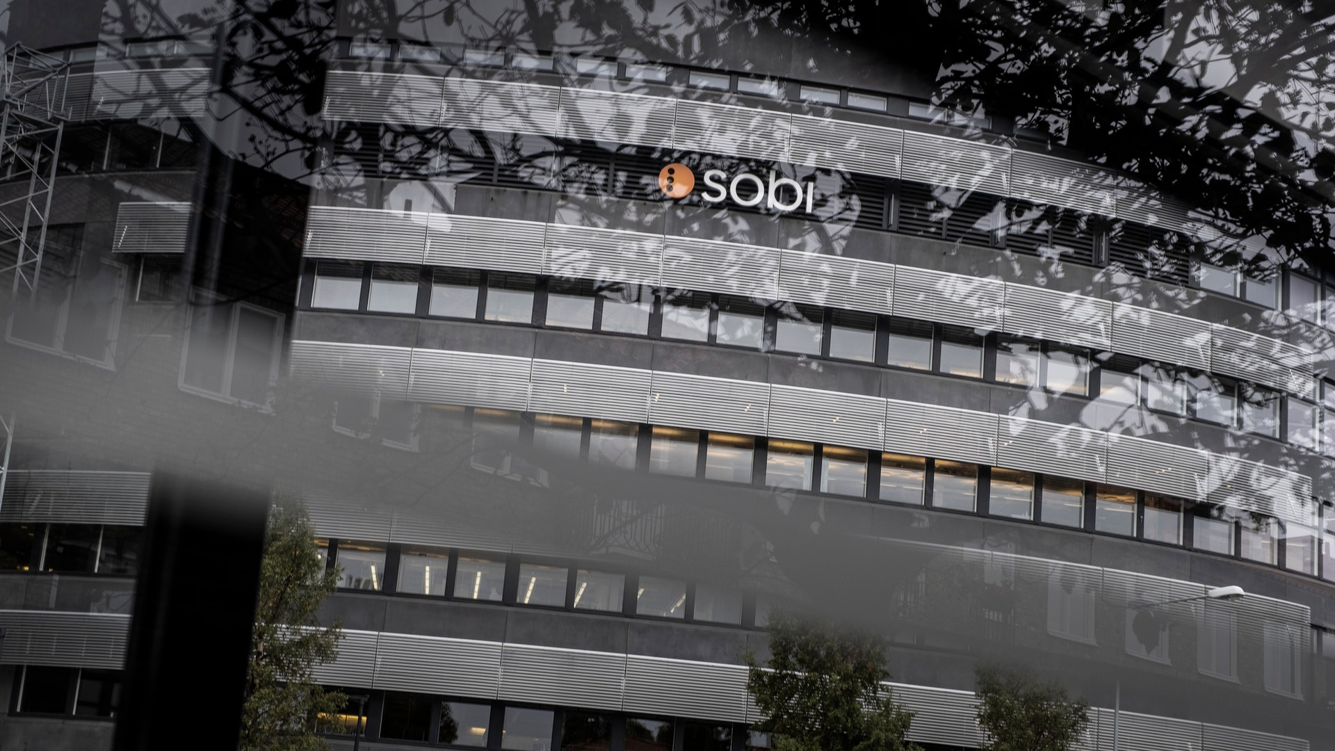 Modernt kontorsbyggnad med SOBI-logotyp på fasaden. Byggnaden har horisontella fönsterband och metallisk-glasarkitektur. Gröna växter vid byggnadens bas. Fotot är taget från låg vinkel uppåt mot himlen.